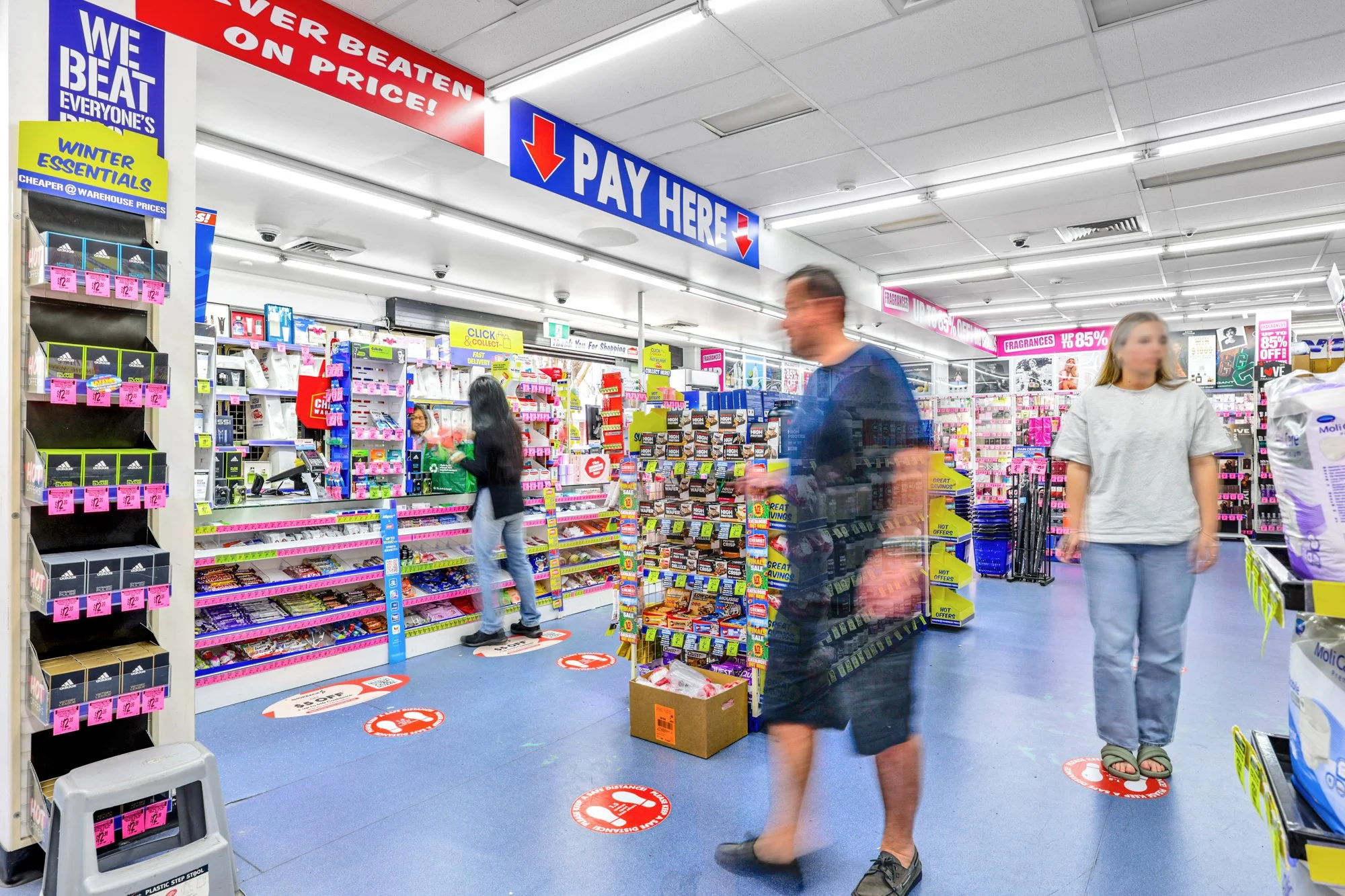 Interior of a retail store with customers standing on social distancing markers, signs indicating 'Pay Here,' and shelves stocked with various products including snacks and personal care items.