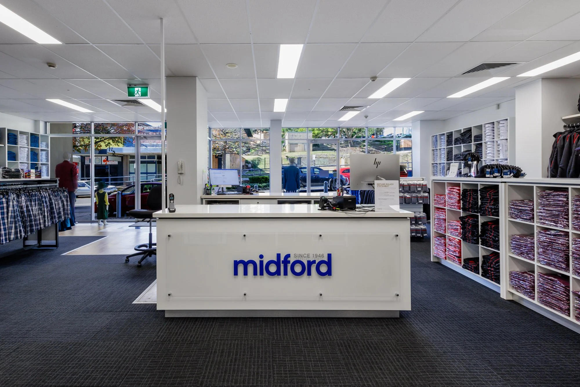Interior of a retail clothing store with a white checkout counter labeled 'midford' and shelves of folded shirts and clothing, large windows in the background showing parked cars and trees.