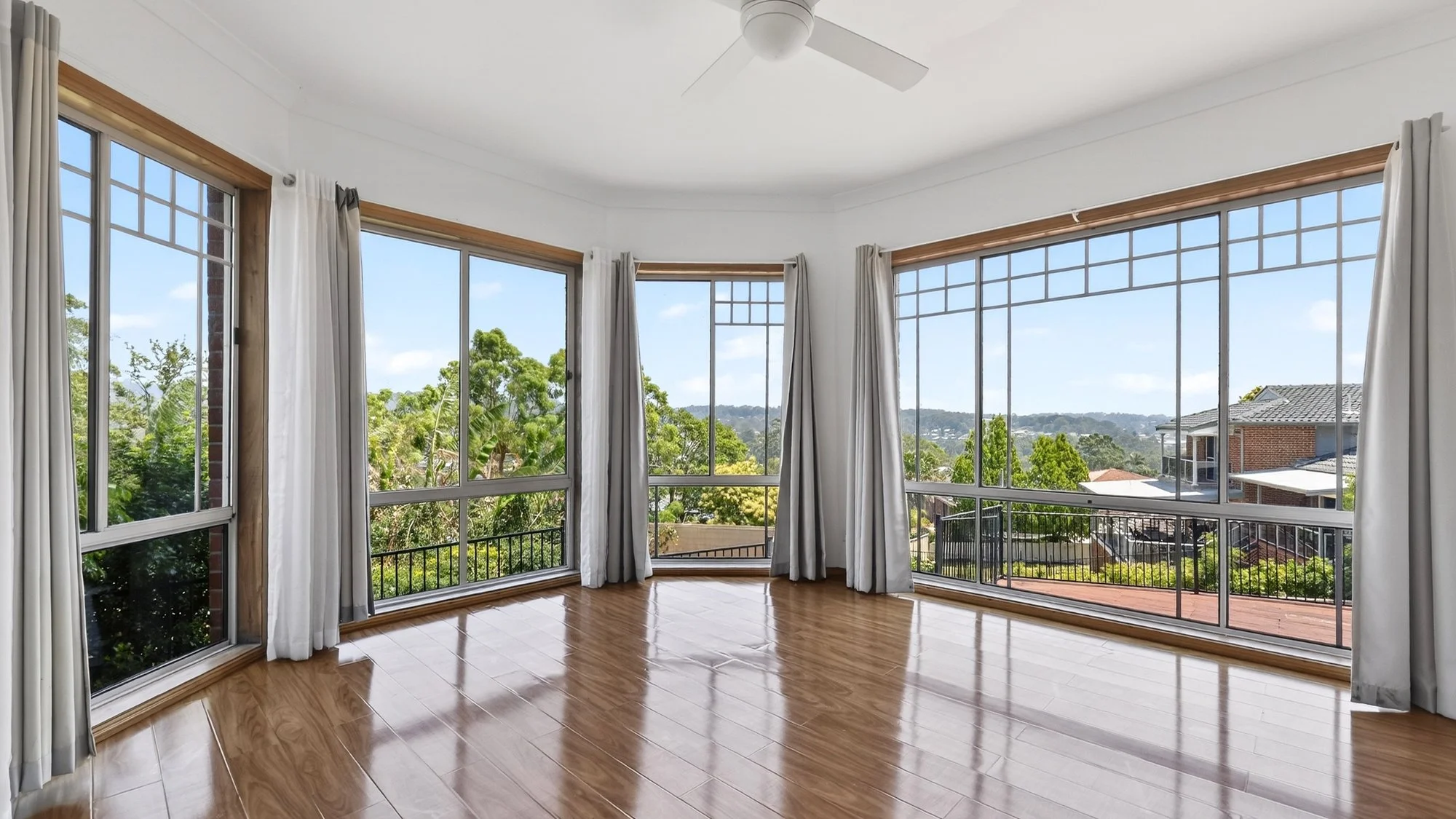 Bright living room with large windows, white curtains, wooden floors, and a view of trees and neighboring houses.