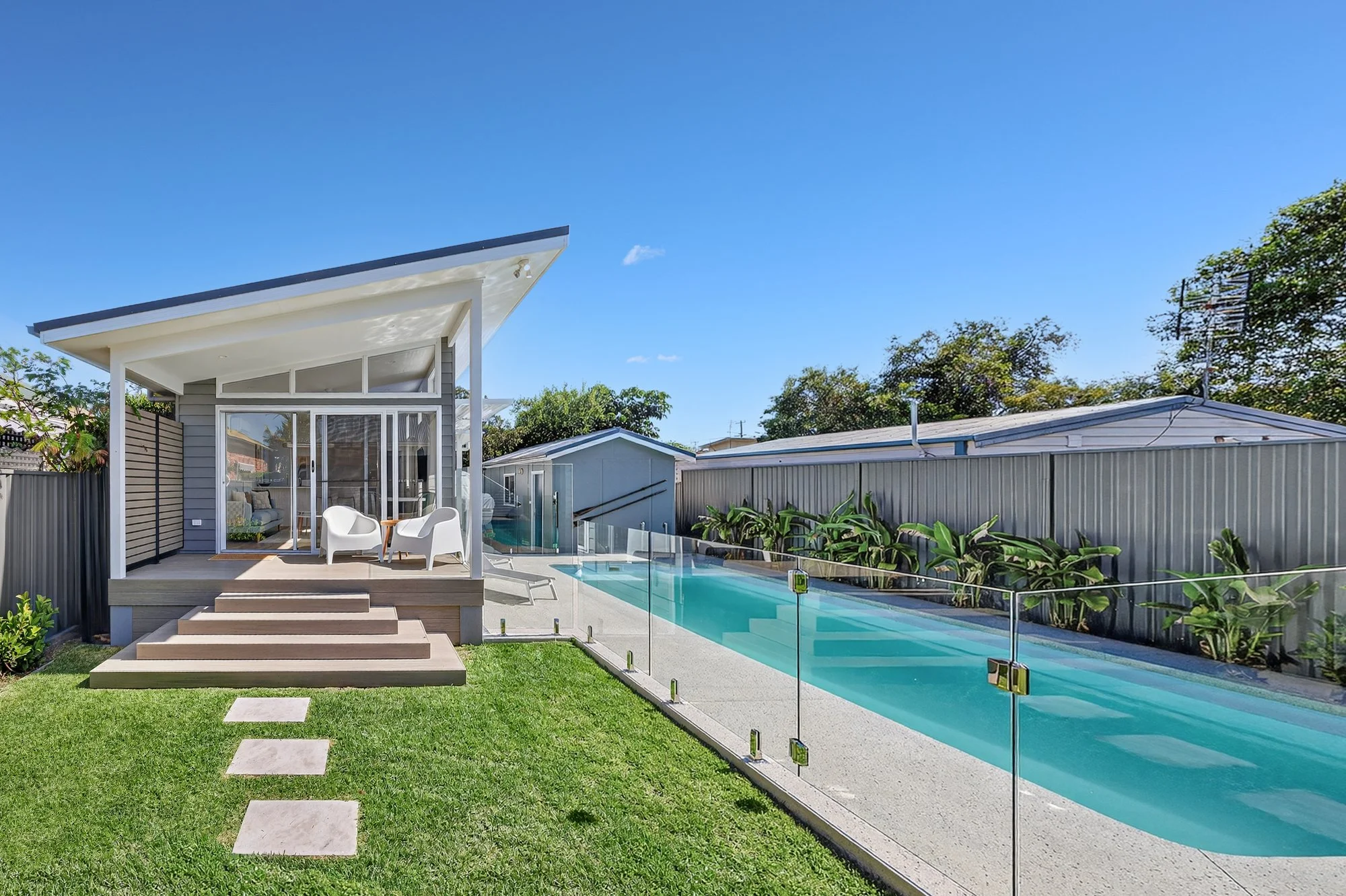 A modern backyard with a small house, outdoor seating area, a narrow swimming pool, and well-maintained green lawn under a clear blue sky.