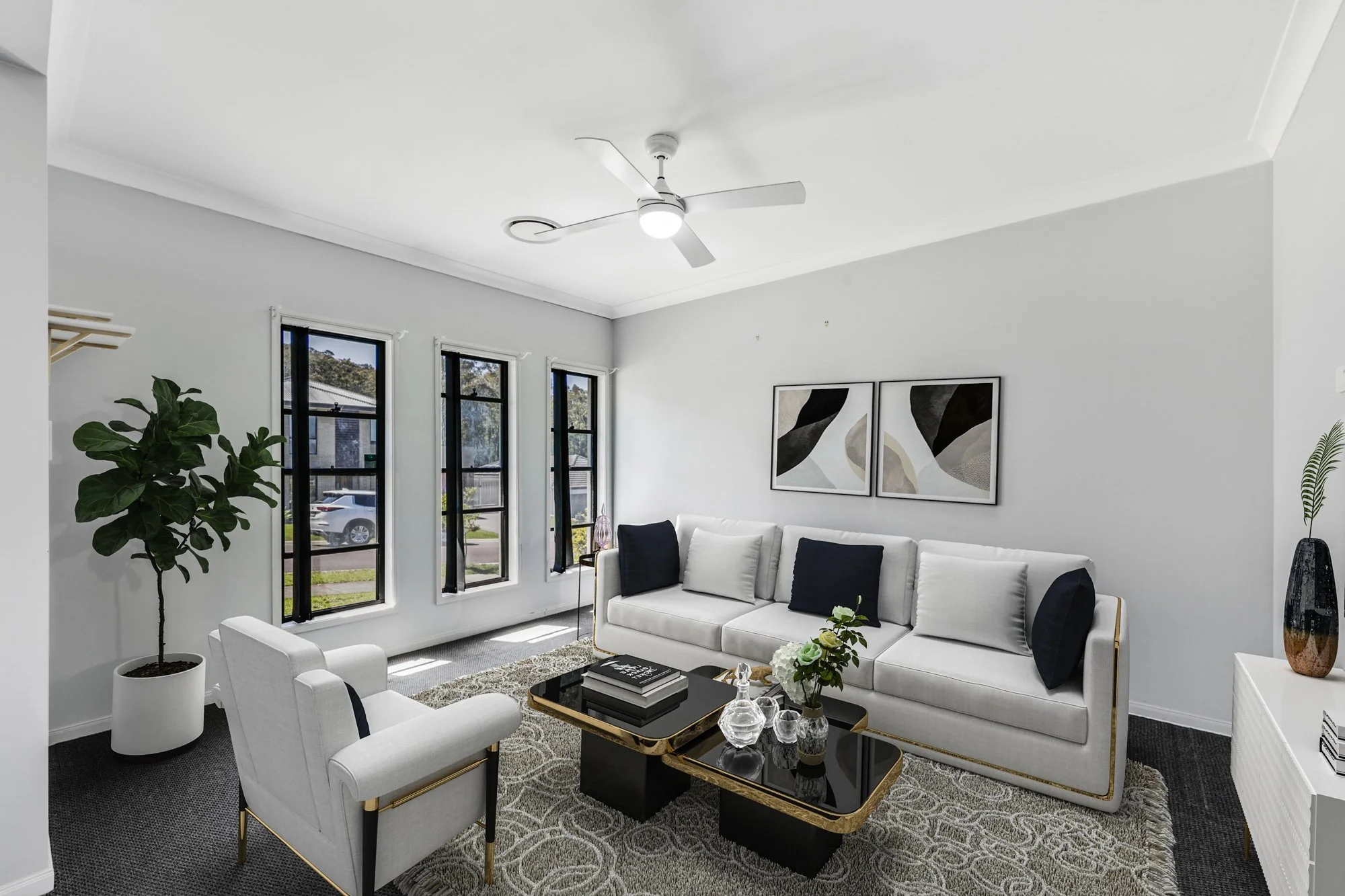 Modern living room with white sofa, black and white pillows, black coffee tables with gold edges, armchair, potted plant, framed artwork, and large windows with black blinds.
