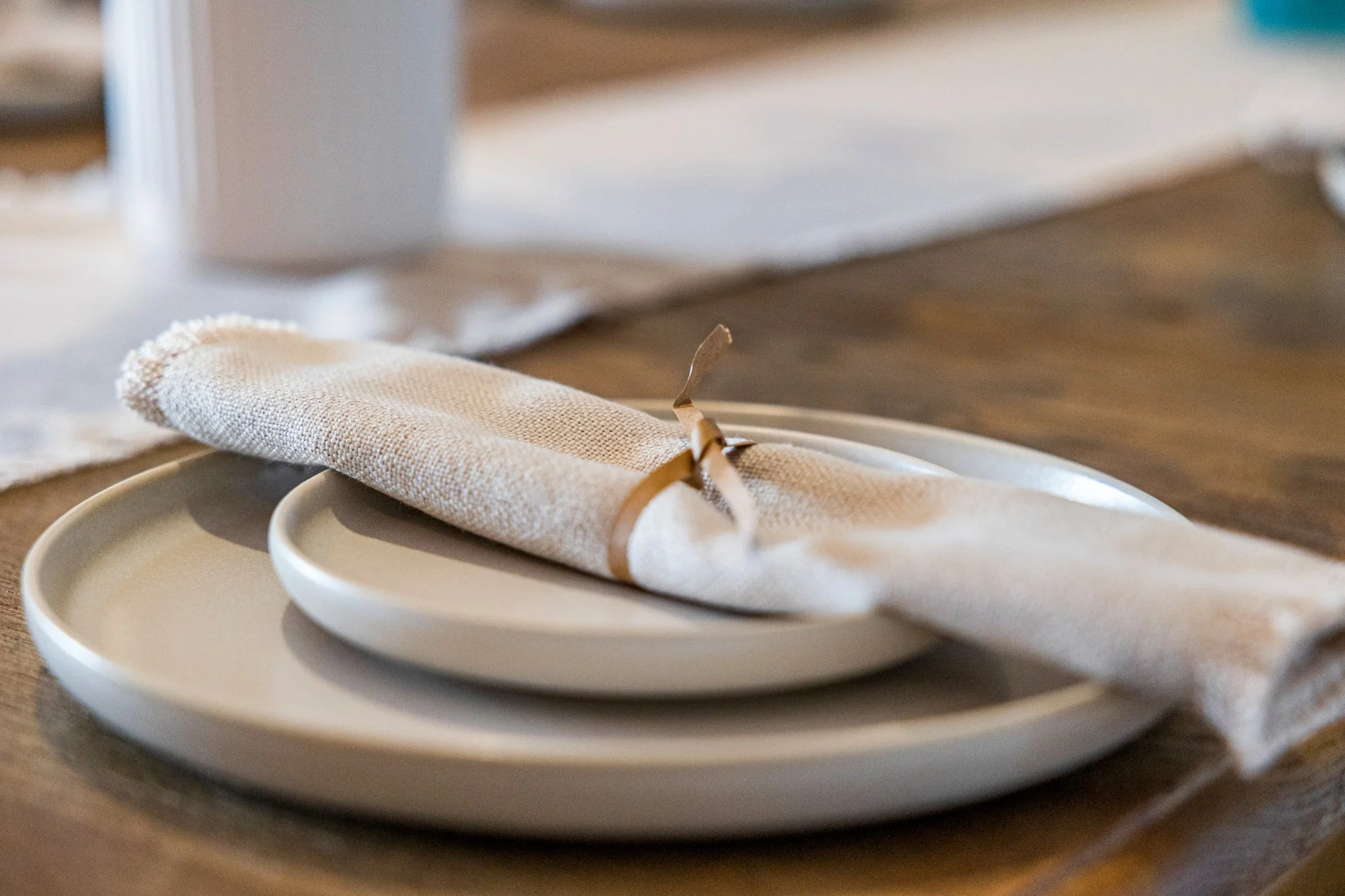 A beige cloth napkin wrapped around a white plate with a smaller matching plate inside, on a wooden table, secured with a tan napkin ring.