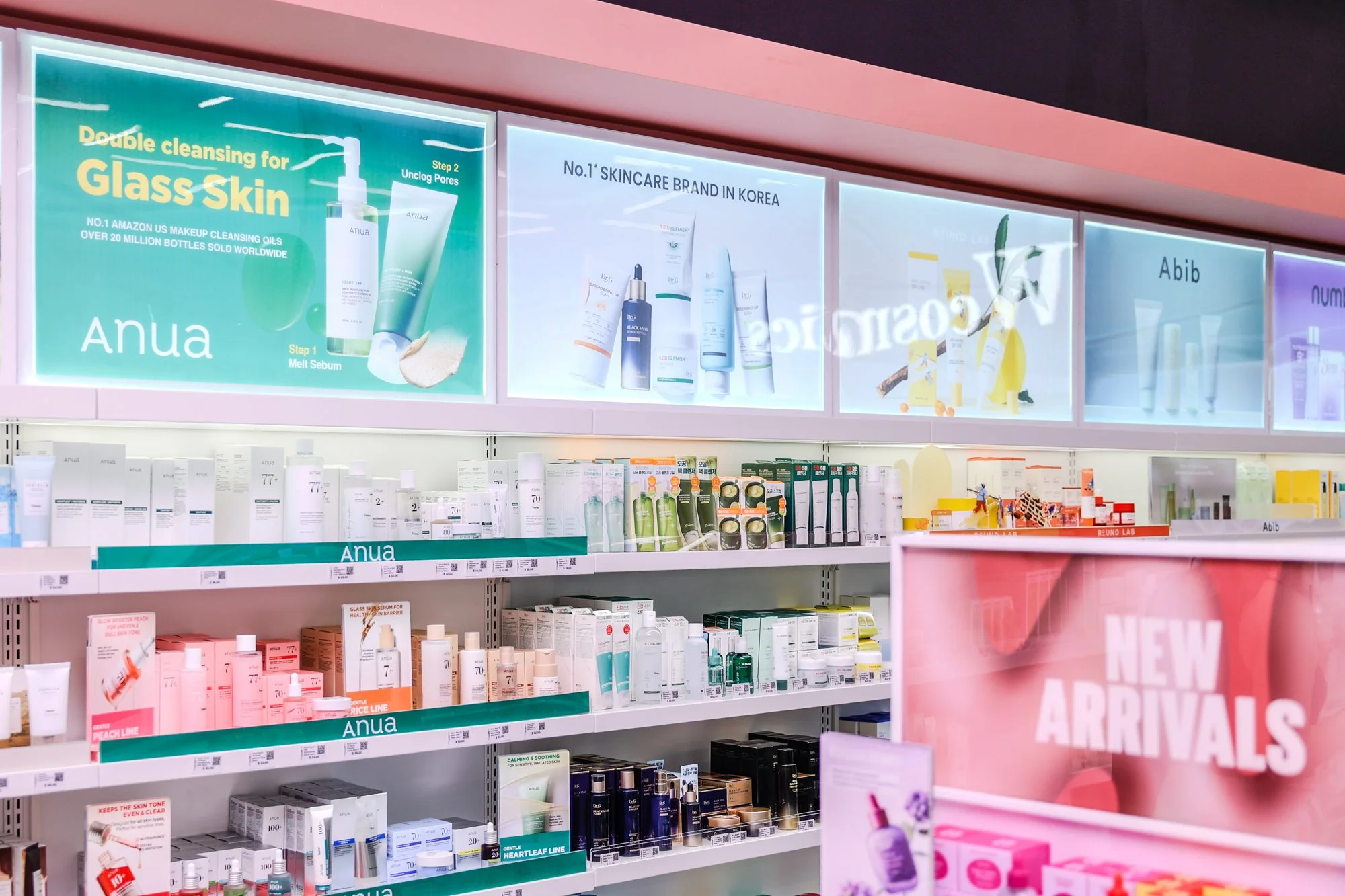 Shelves displaying skincare products at a store, with promotional signs and advertisements for brands like Anua, Abib, and Round Lab, and a pink sign reading 'New Arrivals' in the foreground.