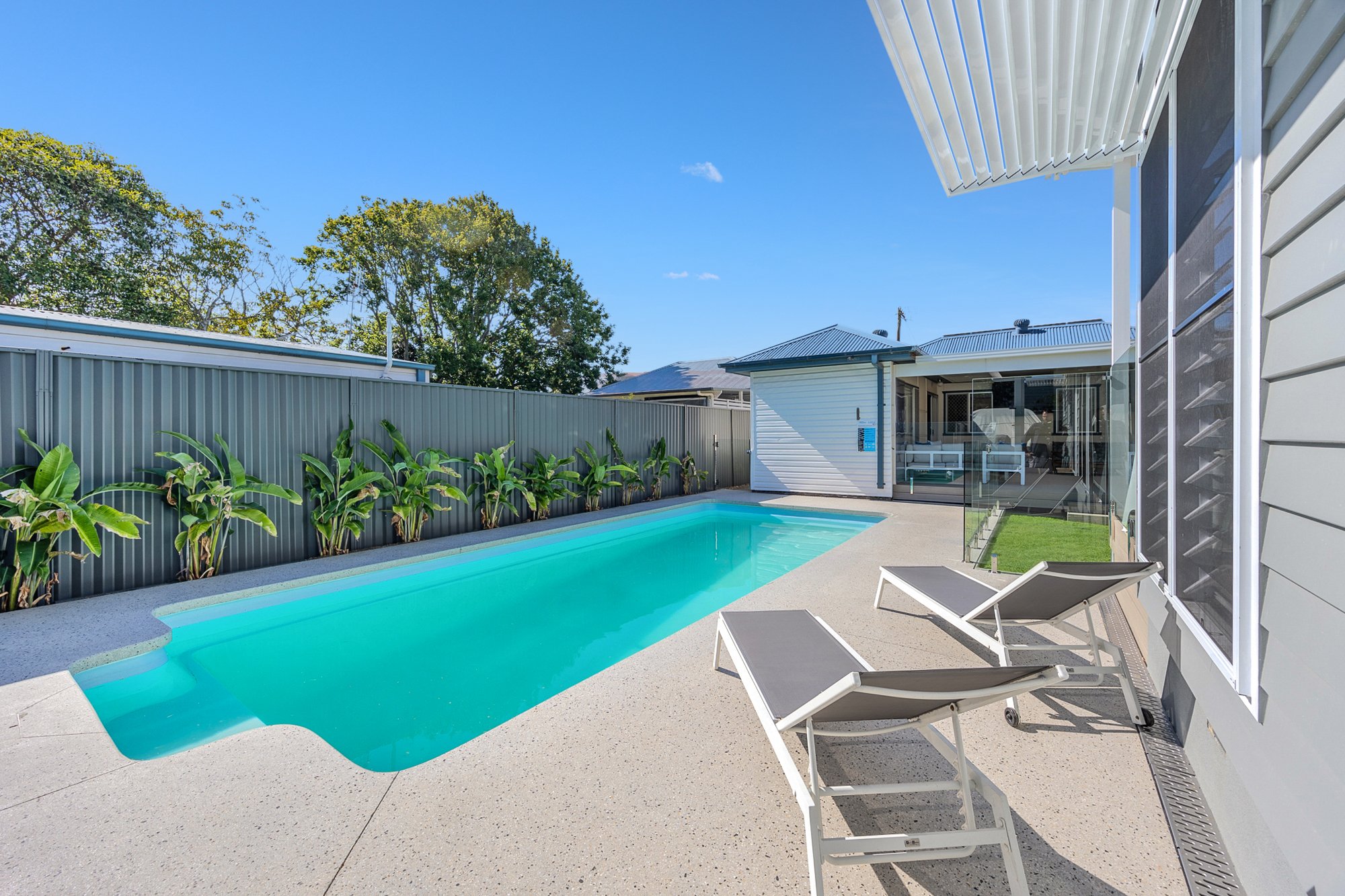 Private backyard with a swimming pool, two lounge chairs, a fence with plants, and a house with a glass door and window under a clear blue sky.