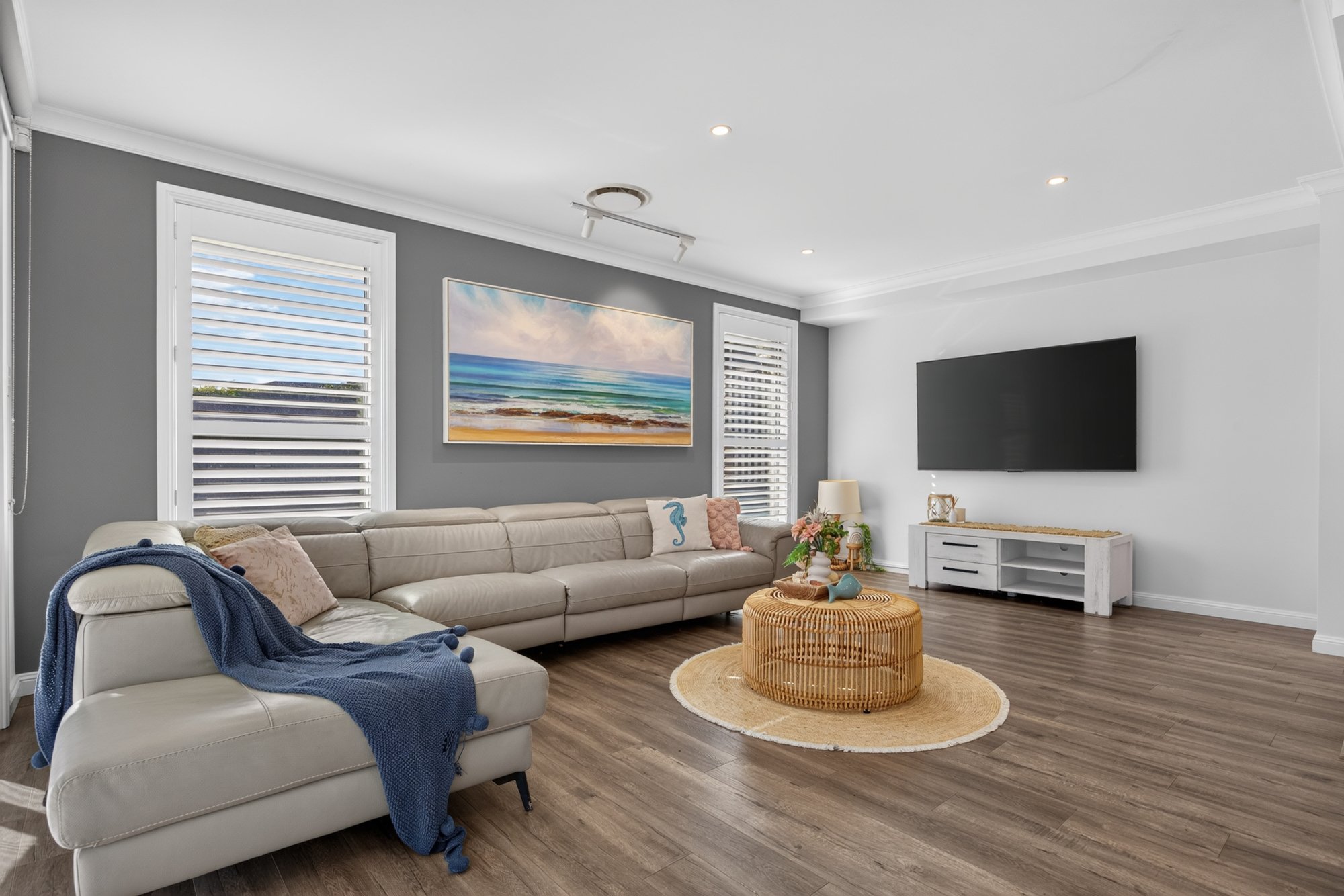 Modern living room with gray accent wall, white couch with light-colored pillows and throw blanket, round wicker coffee table, white TV stand, large flat-screen TV, ocean landscape artwork, white window shutters, hardwood flooring, and soft ceiling l