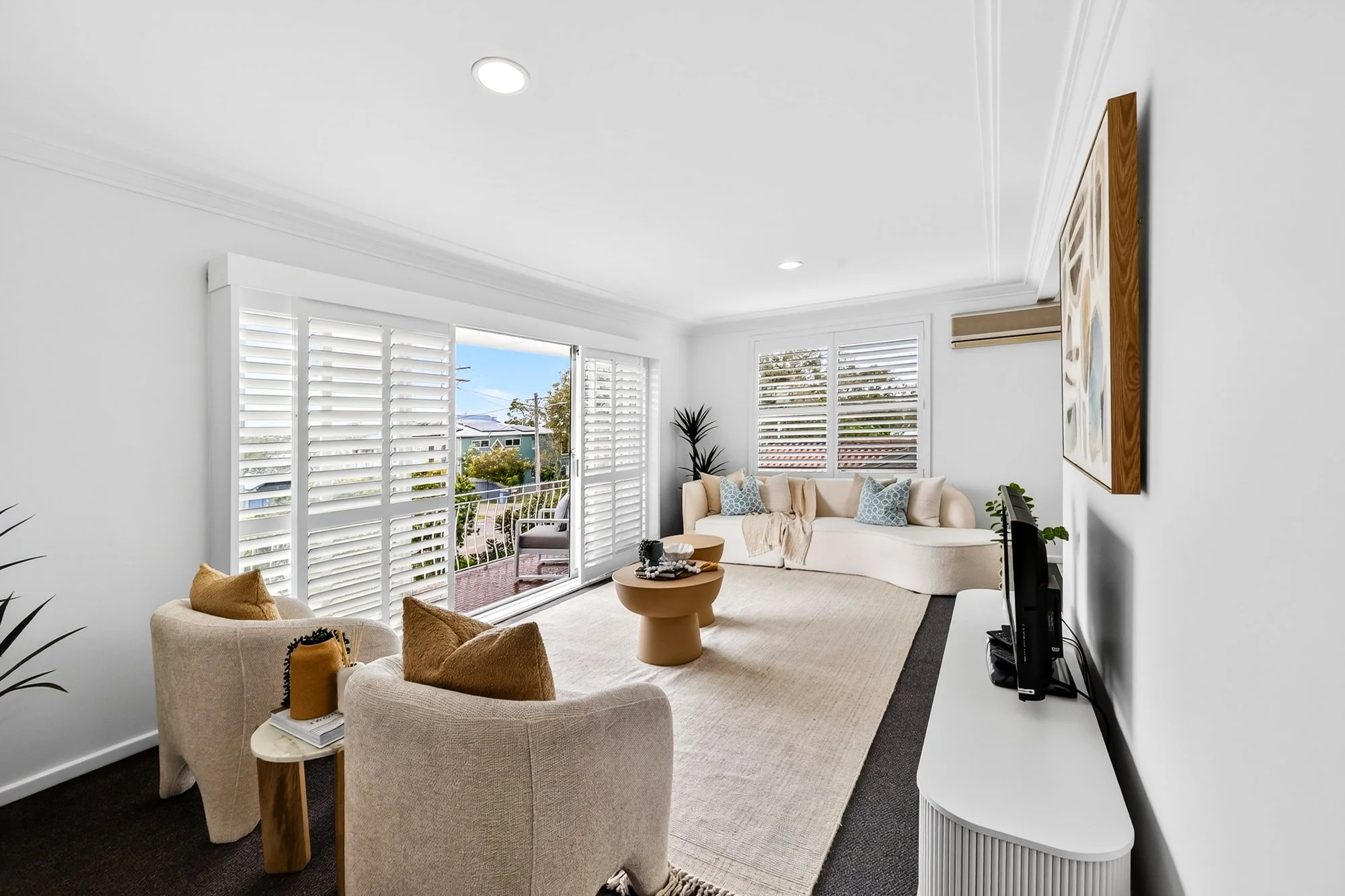 Living room with white walls, large windows with white shutters, beige sectional sofa with blue and beige pillows, two beige armchairs with brown pillows, a beige area rug, a round beige coffee table, a white media console with a TV, a painting on the wall, and a door leading to a balcony with outdoor seating.