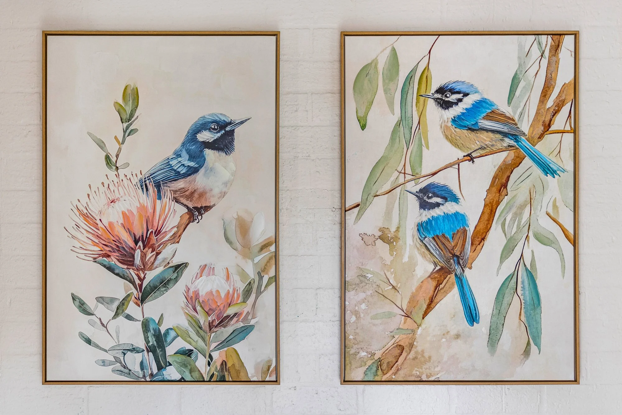 Two watercolor paintings of blue and white birds perched on branches with green leaves, framed and hanging on a white brick wall.