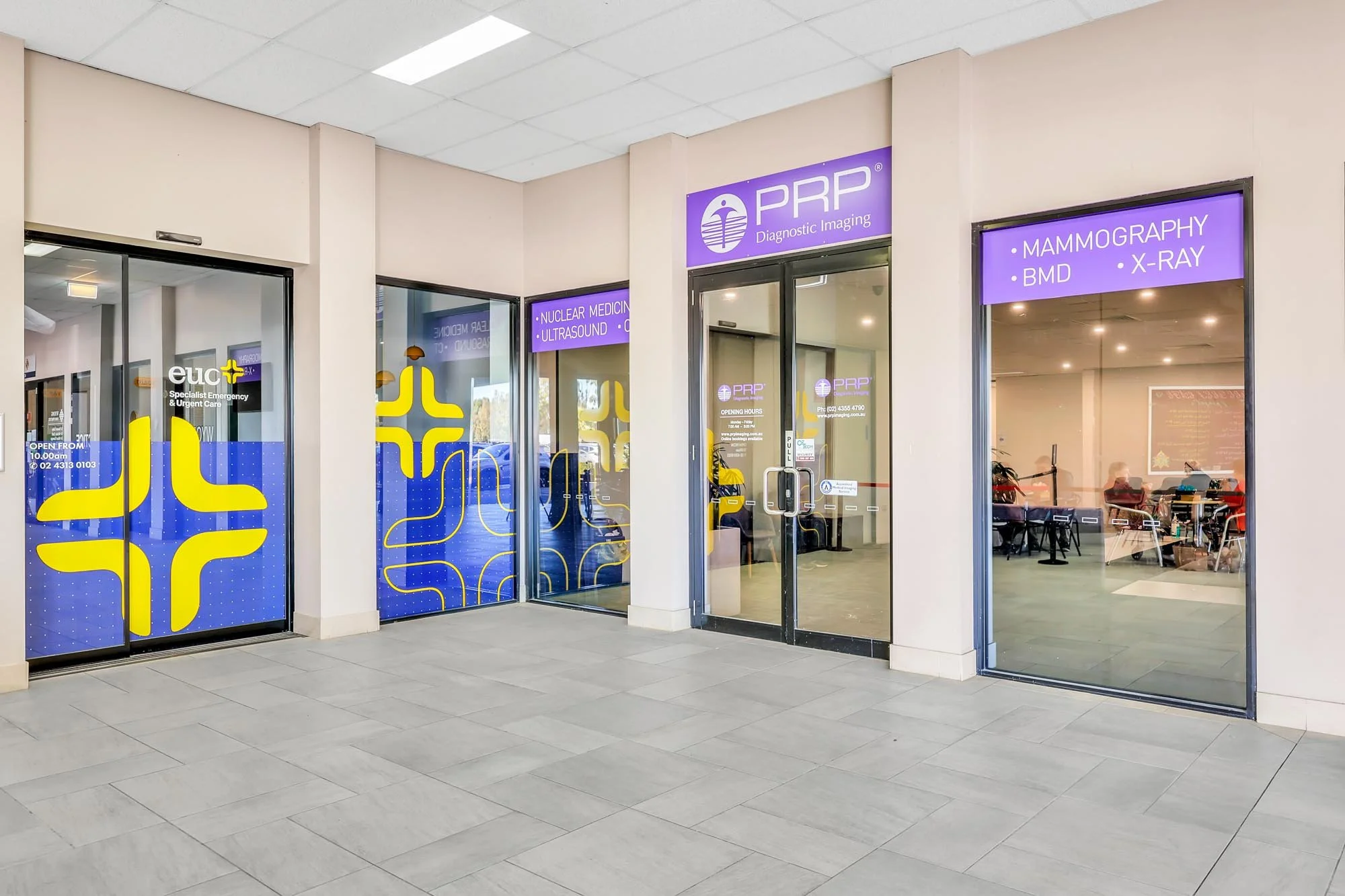 Exterior view of a medical imaging clinic with glass doors and windows, purple signage featuring a medical symbol, and a waiting area visible inside with people seated.