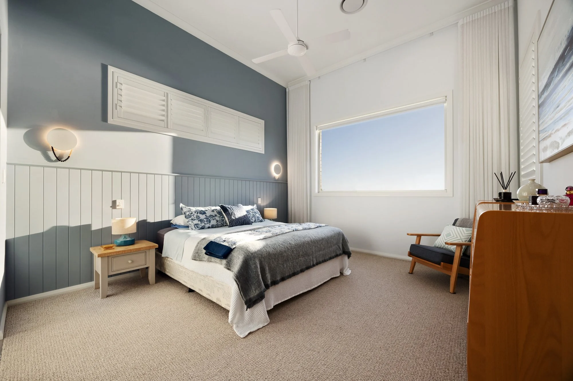 A bedroom with a bed, side table, and a chair near a window with white shutters. The room has light walls, a ceiling fan, and lamps on either side of the bed.