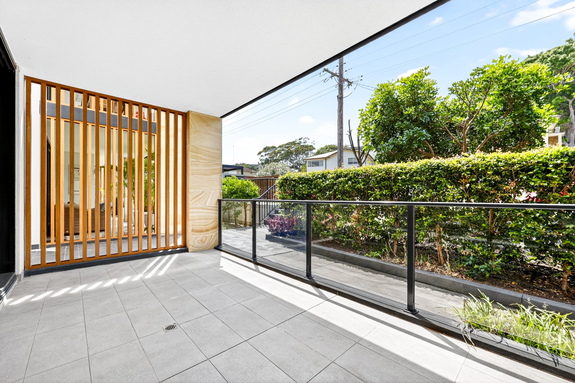 Outdoor balcony with gray tiled floor, black metal railing, potted plants, lush green bushes and trees, and a partly cloudy sky.