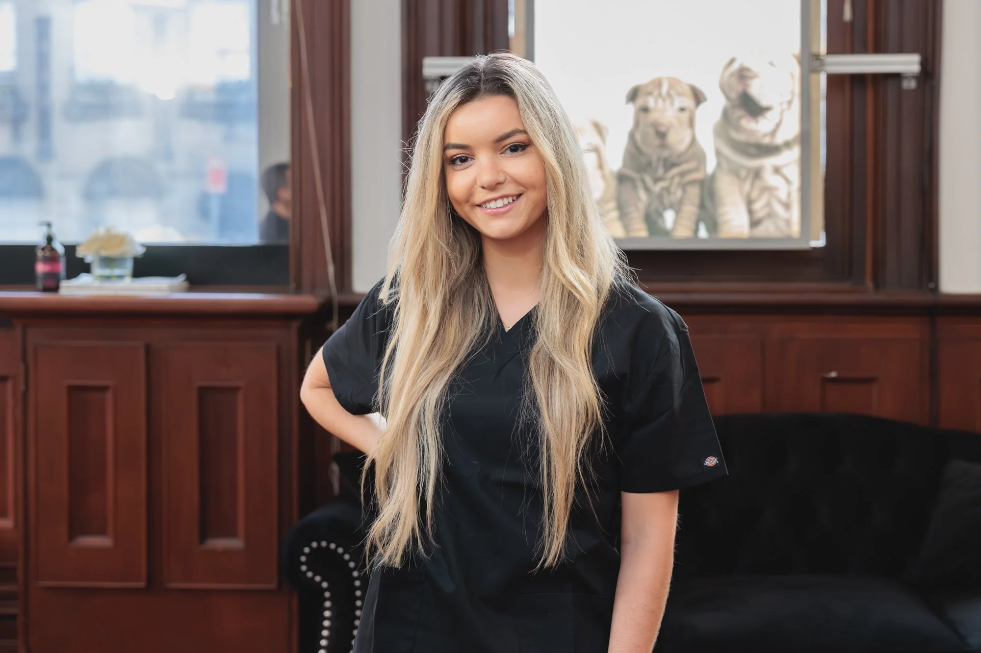 Young woman with long blonde hair smiling in a black uniform inside a room with wood-paneled walls and tiger artwork in the background.