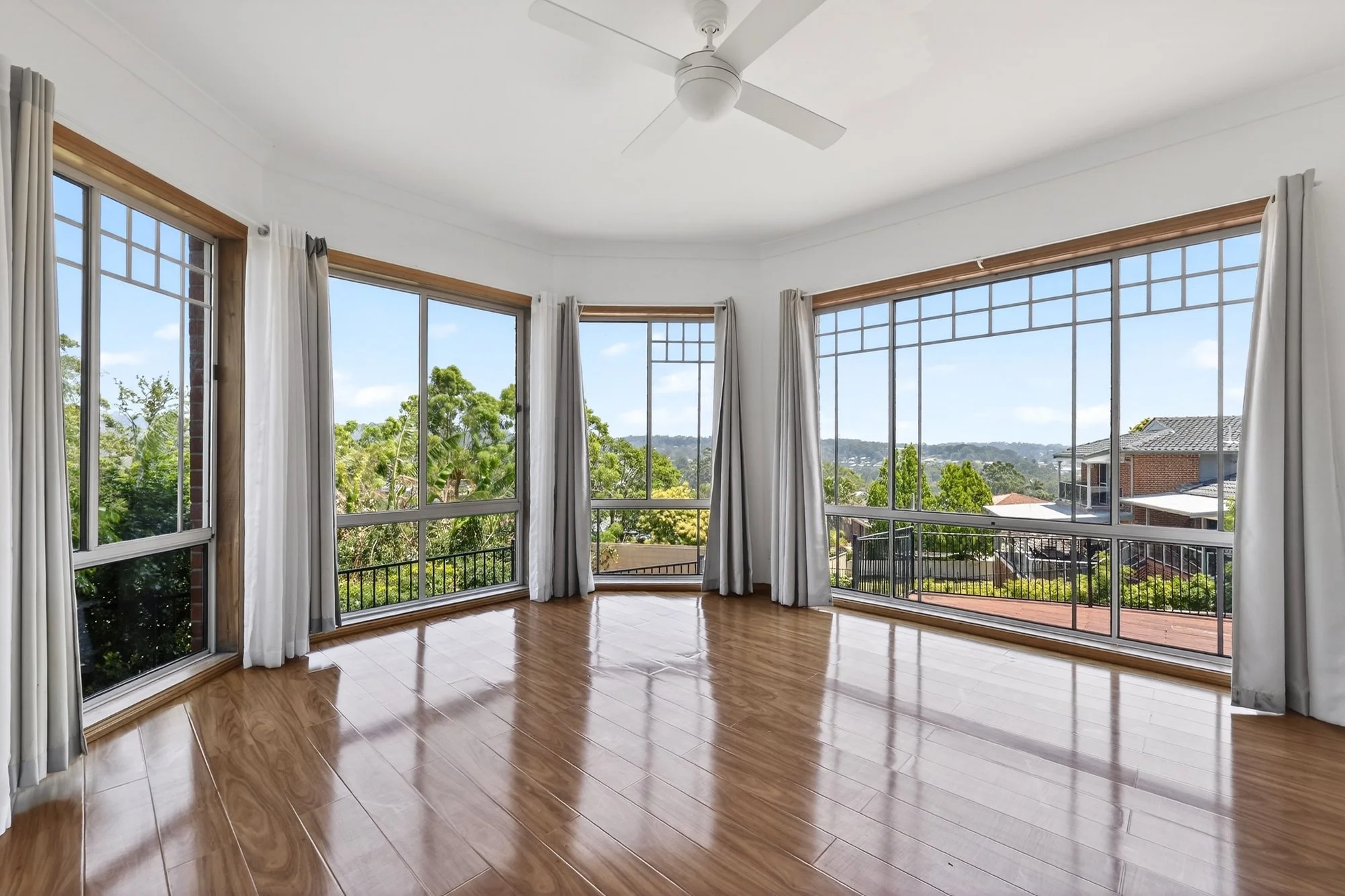 Empty room with large windows, white walls, wooden floors, white curtains, and a ceiling fan, with a view of trees and neighboring houses outside.