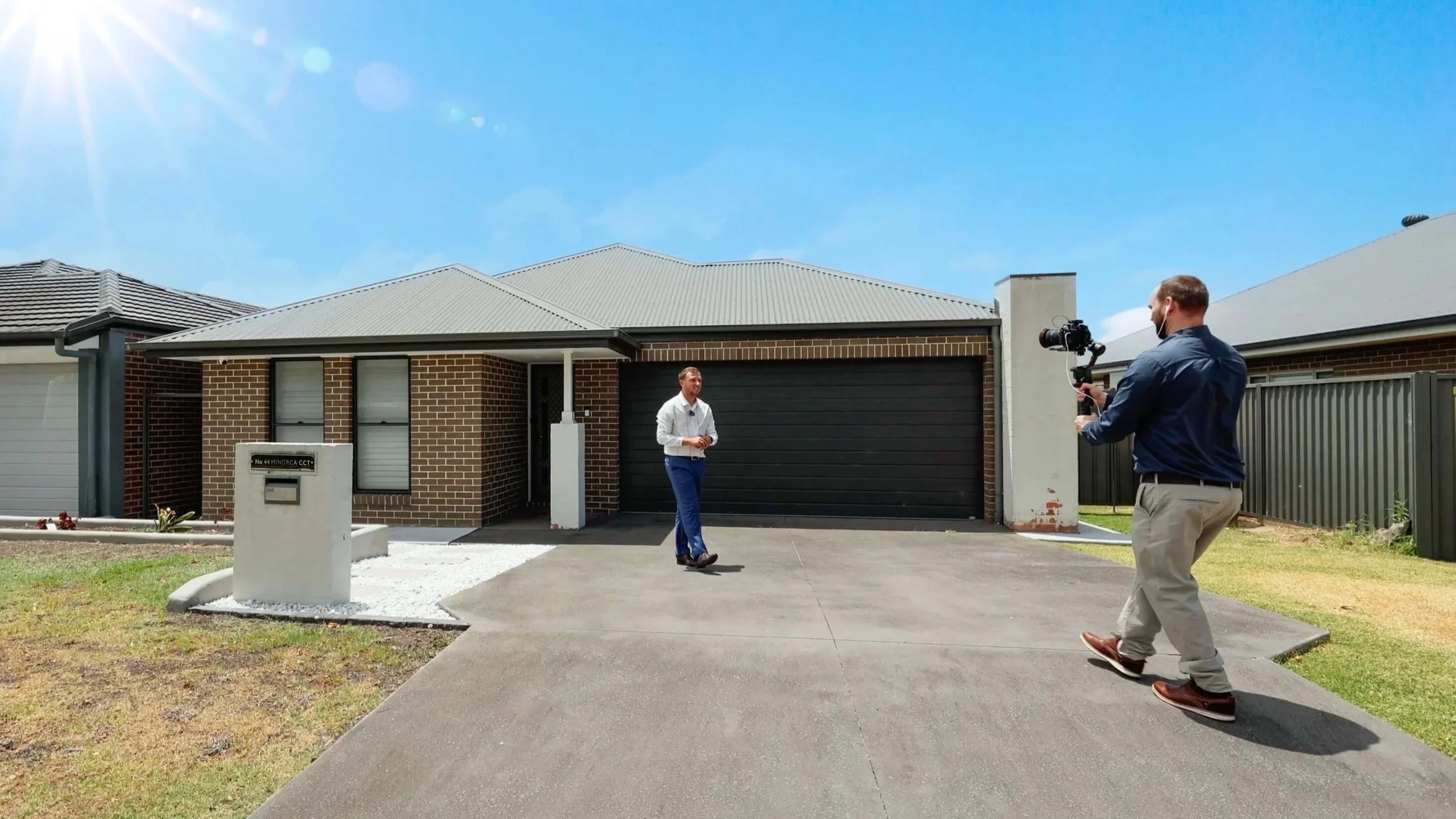 A man in a white shirt and blue pants being filmed by a cameraman in front of a brick house with a black garage door on a sunny day.