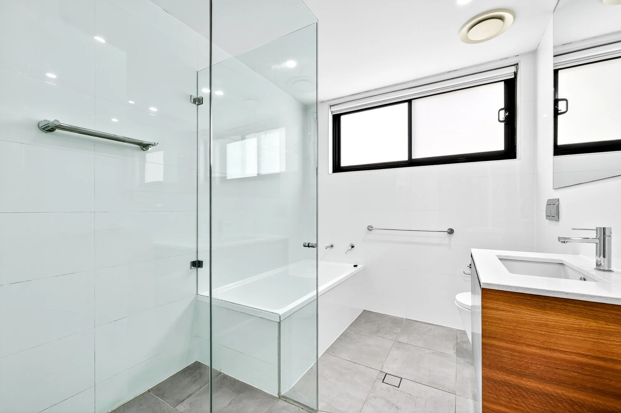 Modern bathroom with a glass shower enclosure, a white bathtub, a window with black frames, a toilet, and a wooden vanity with a white sink.
