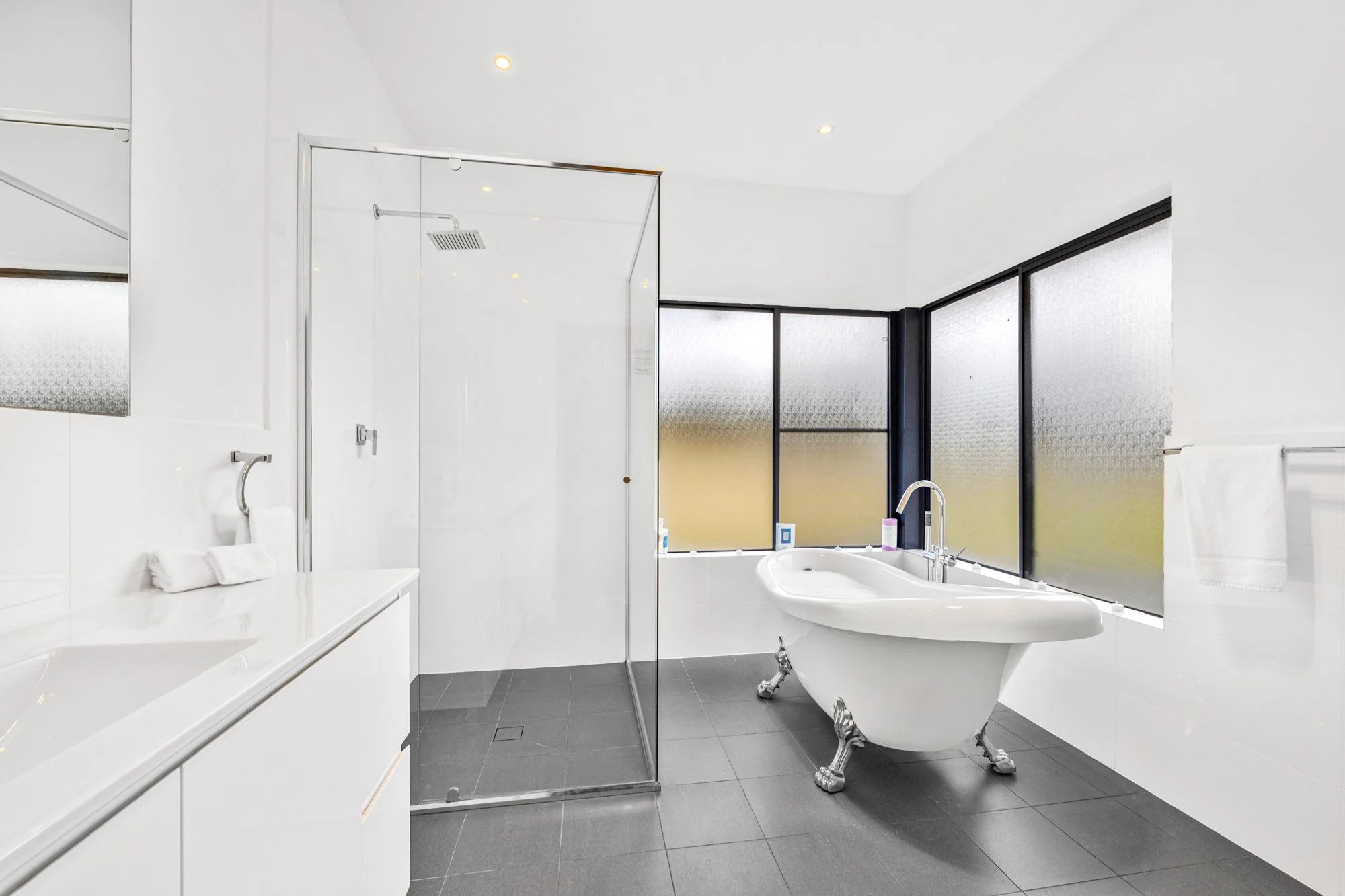 Modern bathroom with a clawfoot bathtub, frosted sliding windows, a glass shower enclosure, a white vanity with towels, and dark tiled flooring.