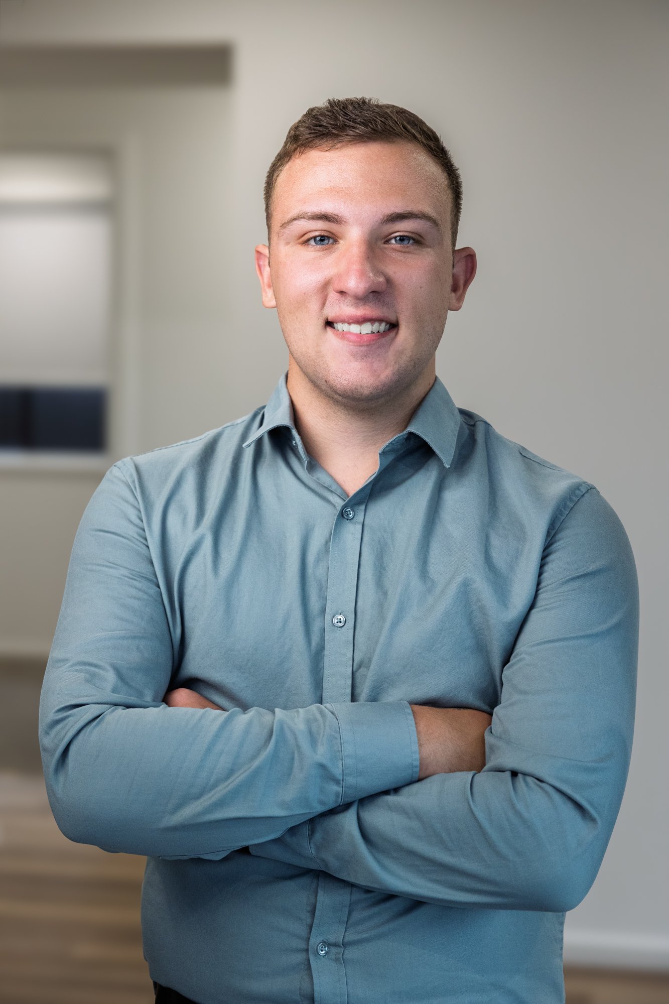 A portrait of a young man with short brown hair and blue eyes, smiling, wearing a blue button-up shirt with arms crossed, standing indoors against a neutral background.
