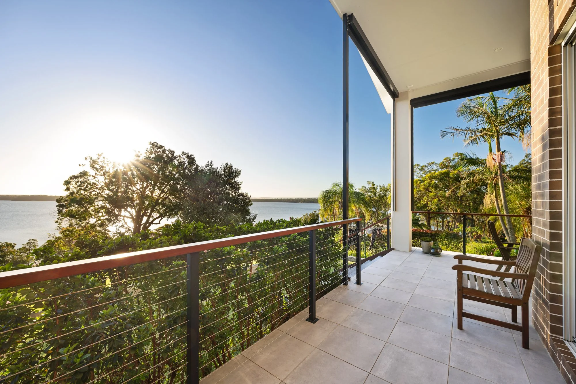 Balcony with a wooden chair, potted plants, and a view of trees, water, and the sunset.
