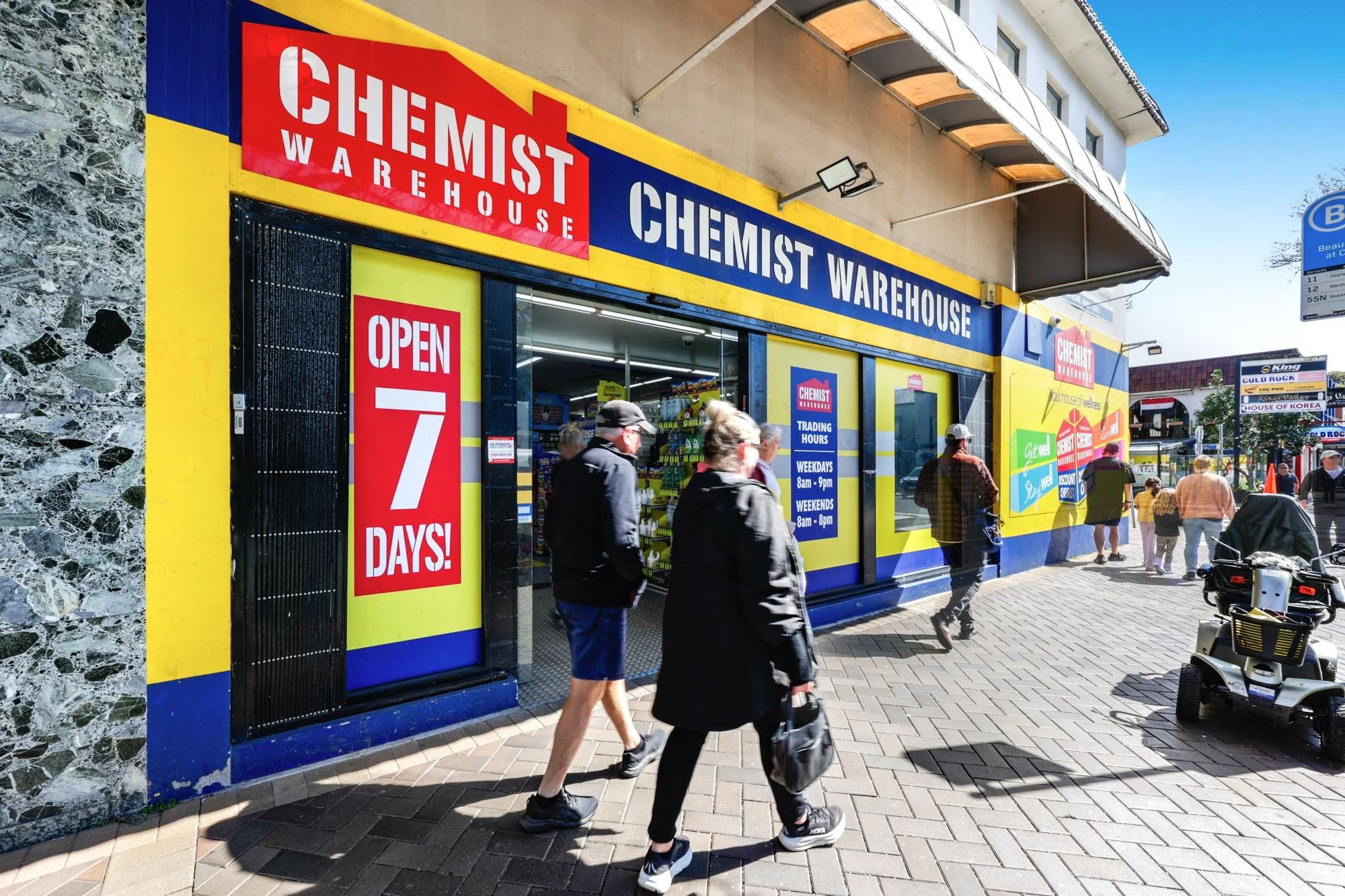 People walking past a Chemist Warehouse store with bright yellow and blue exterior, large signage, and open sign, in a shopping plaza on a sunny day.