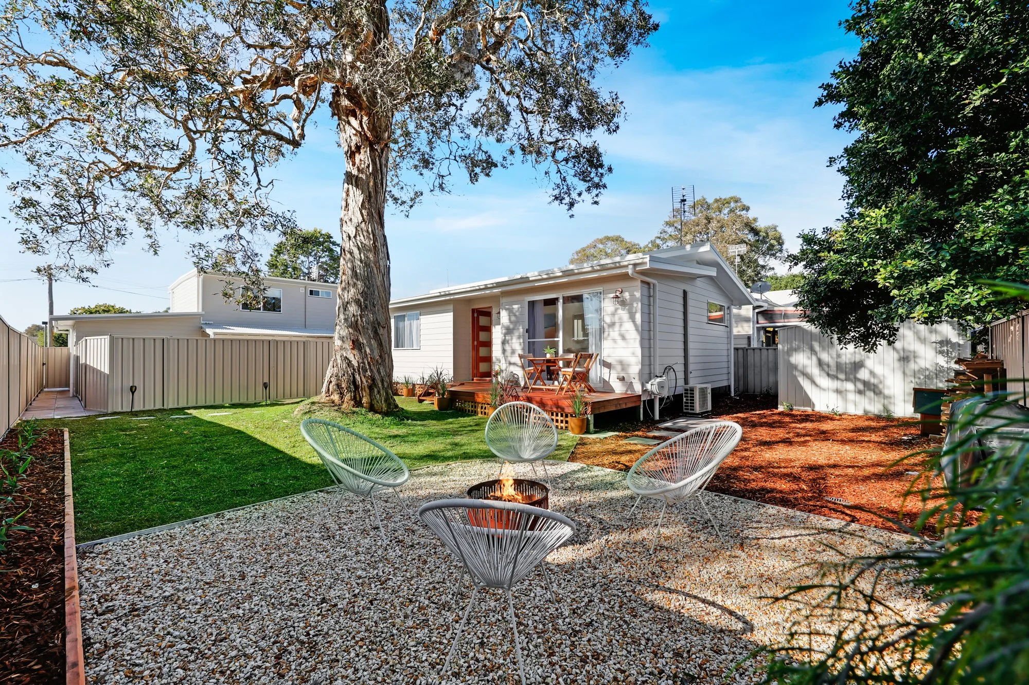 Blue sky, new modern deck, green grass, paperbark tree, while stones, landscaped garden, fire pit, white single story house photographed for Airbnb listing in Budgewoi