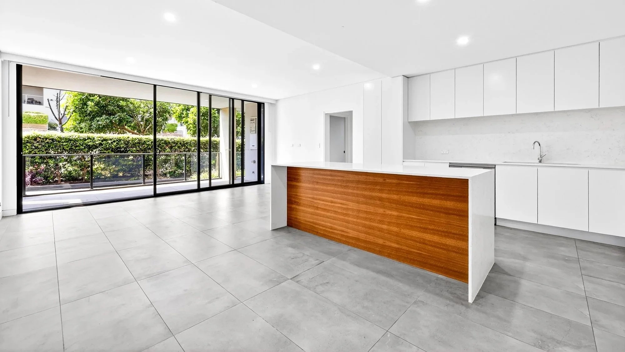 Modern, empty kitchen with white cabinets, a white marble backsplash, and a large island with a wood panel, overlooking a patio with glass sliding doors and greenery outside.