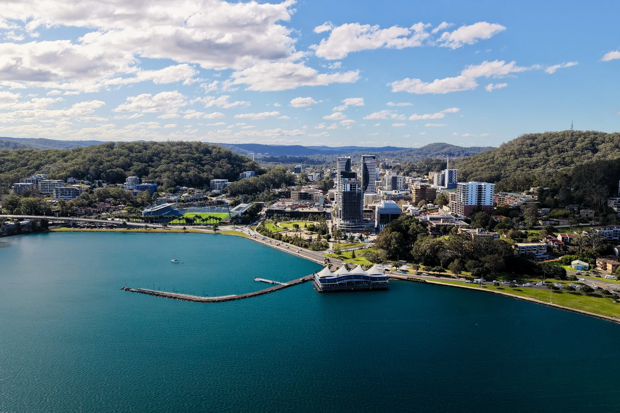 Aerial view of a city with a large body of water in the foreground, surrounded by green hills and modern buildings, under a partly cloudy blue sky.