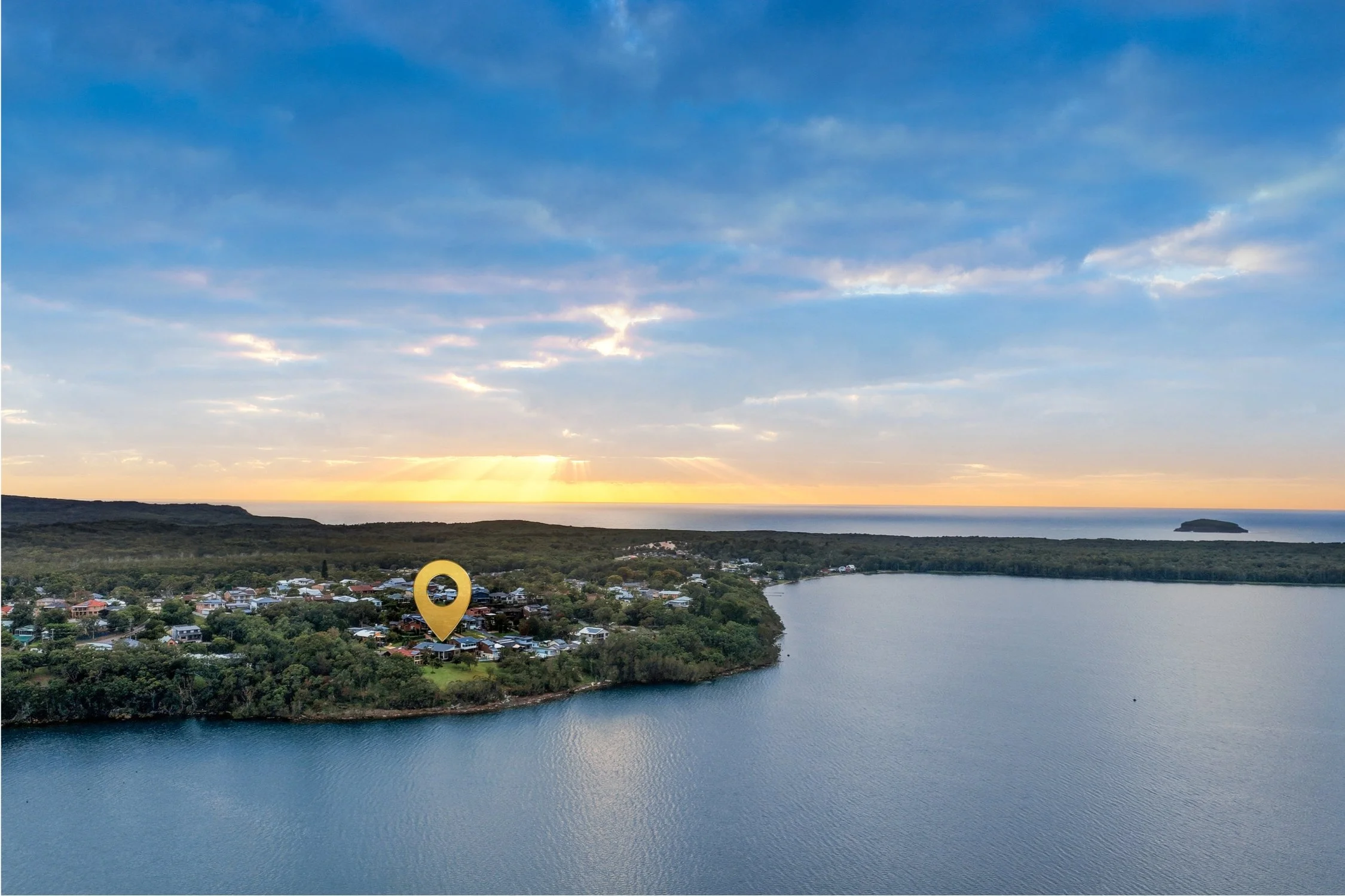 A scenic aerial view of a lakeside town at sunset, with a yellow location marker over a specific building in the town, and a distant island on the horizon.