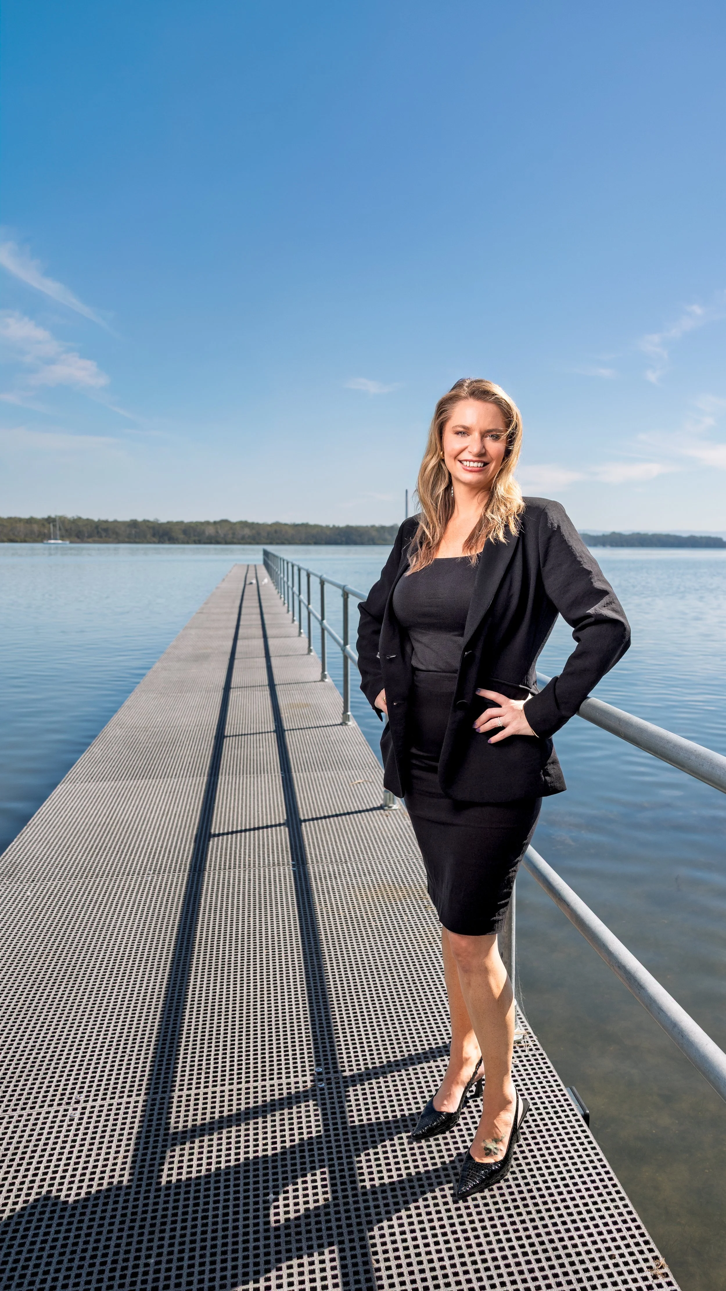 A woman wearing a black dress and blazer standing on a metal dock by a calm body of water, smiling at the camera with her hand on her hip.