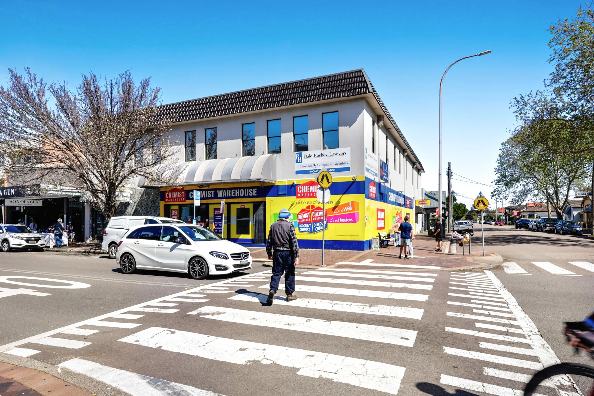 A street scene with a crosswalk, parked cars, a building with colorful advertisements for a hardware store, and pedestrians walking on the sidewalk on a clear, sunny day.