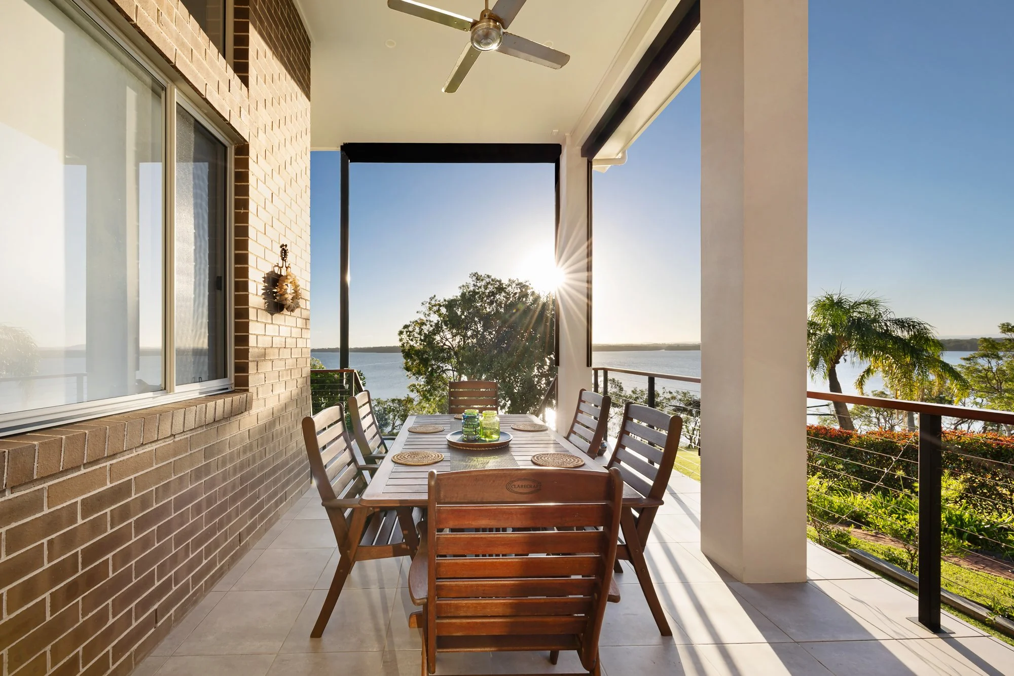 Outdoor dining area on a balcony with a wooden table and chairs, overlooking a lake with trees and a clear sky, sunlight shining.