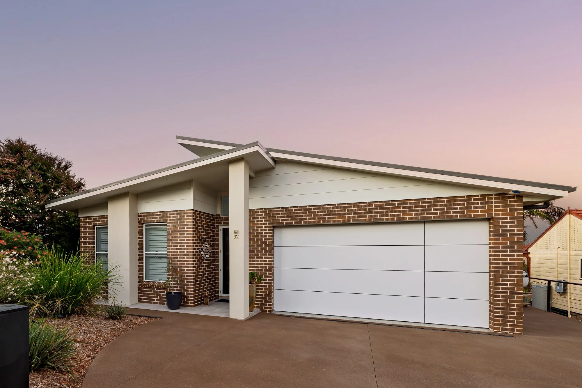 Modern single-story house with brick and white siding exterior, white garage door, small front porch with potted plants, and a curved concrete driveway, during sunset.