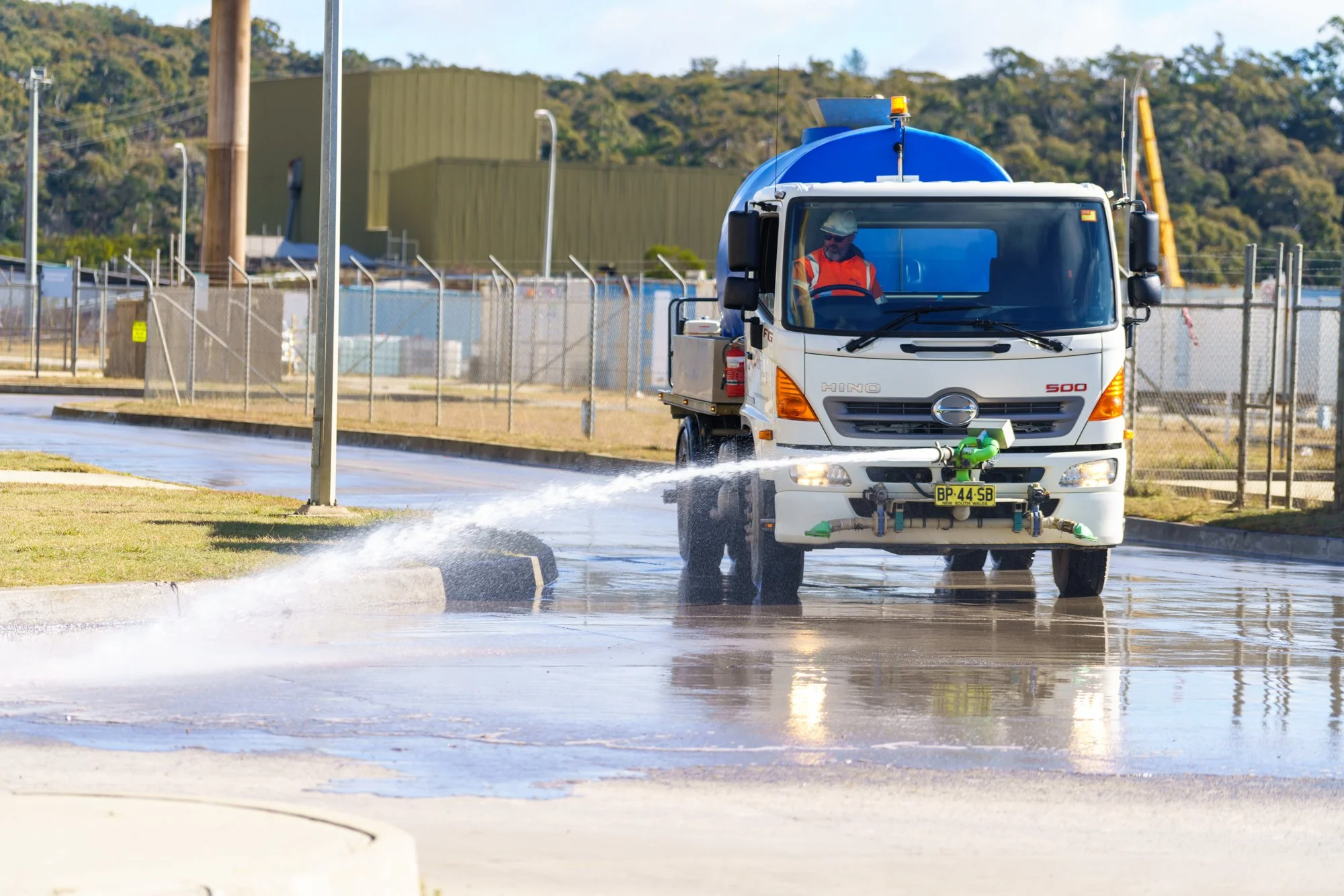 A worker in safety gear disinfects a paved area with a spray truck during daytime.