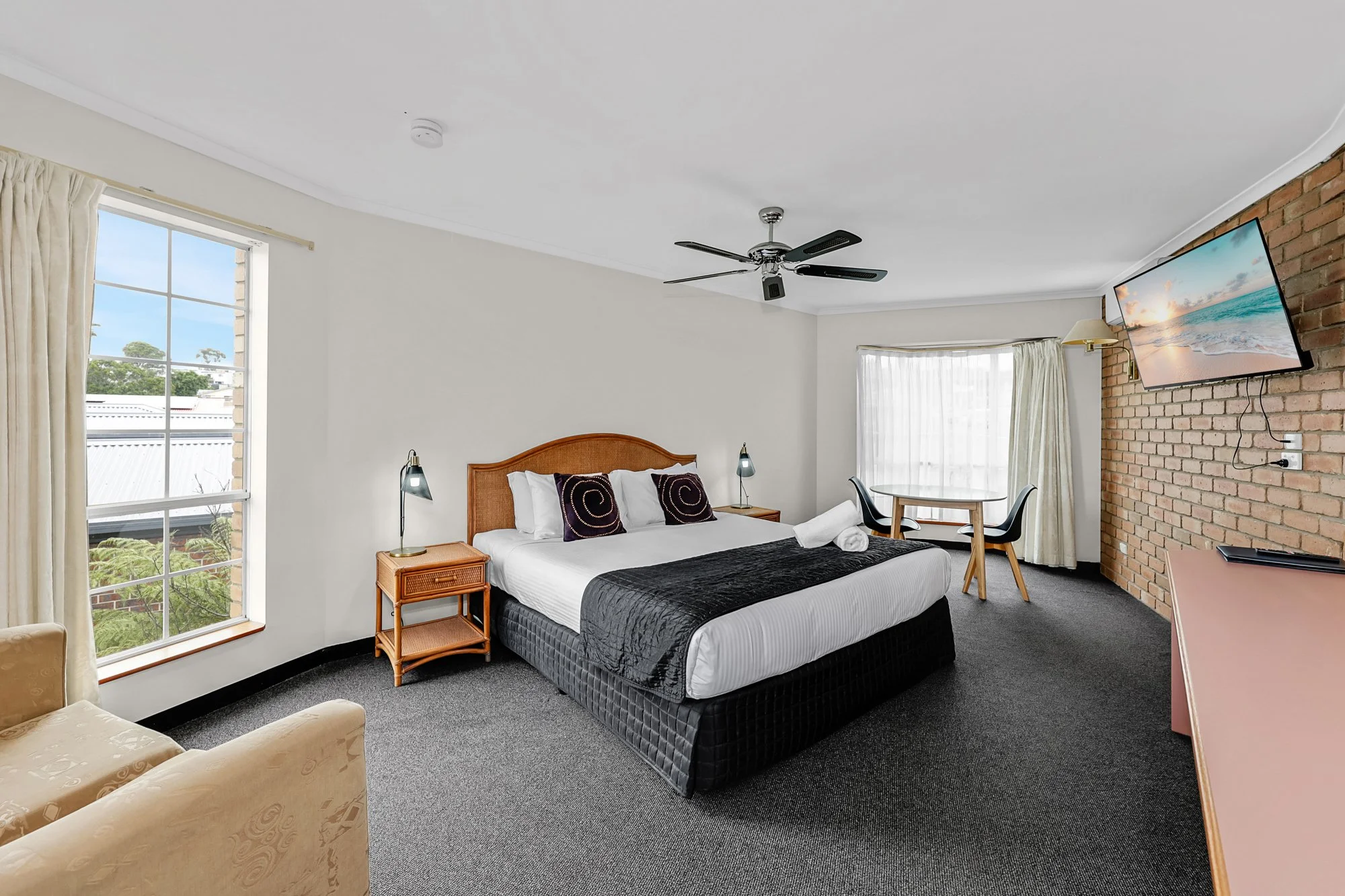 Hotel room with a king-sized bed, decorative pillows, bedside tables with lamps, a ceiling fan, wall-mounted TV, and a small table with chairs near large windows.