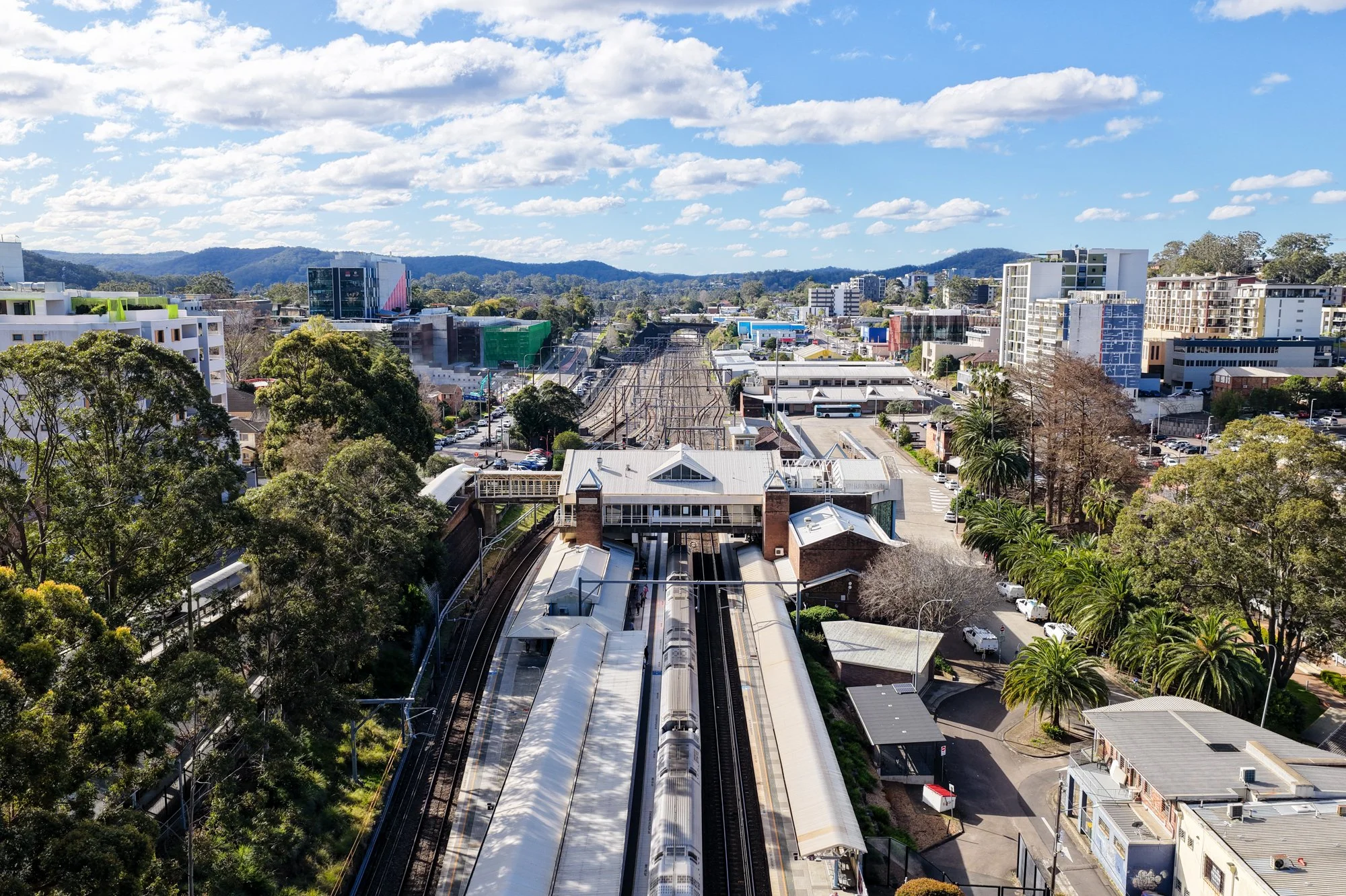 An aerial view of a train station surrounded by trees, buildings, and a parking lot on a sunny day with a blue sky and scattered clouds.