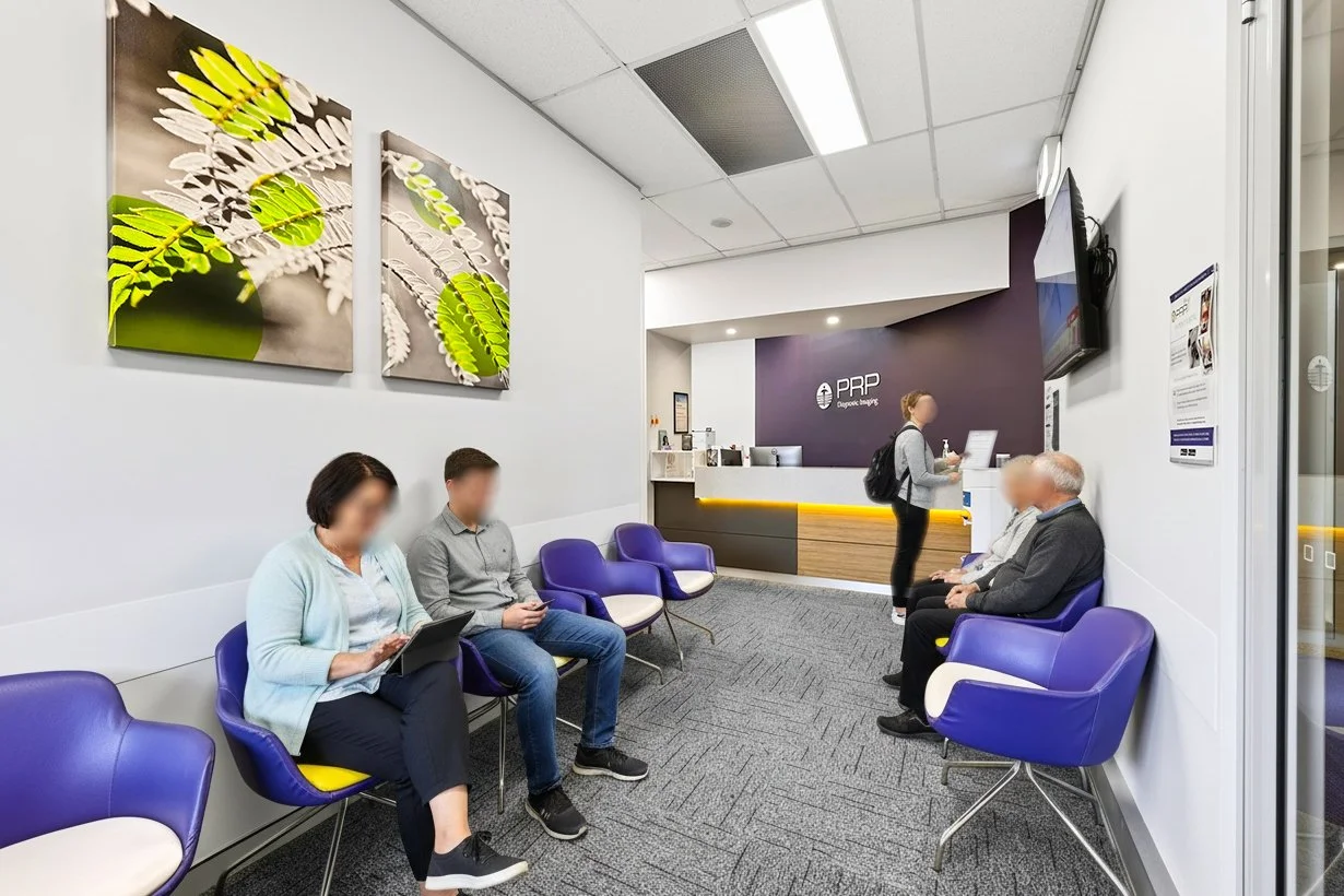 Interior of a medical office waiting room with people sitting, some using devices, purple chairs, wall art, reception desk, and a staff member assisting patients.