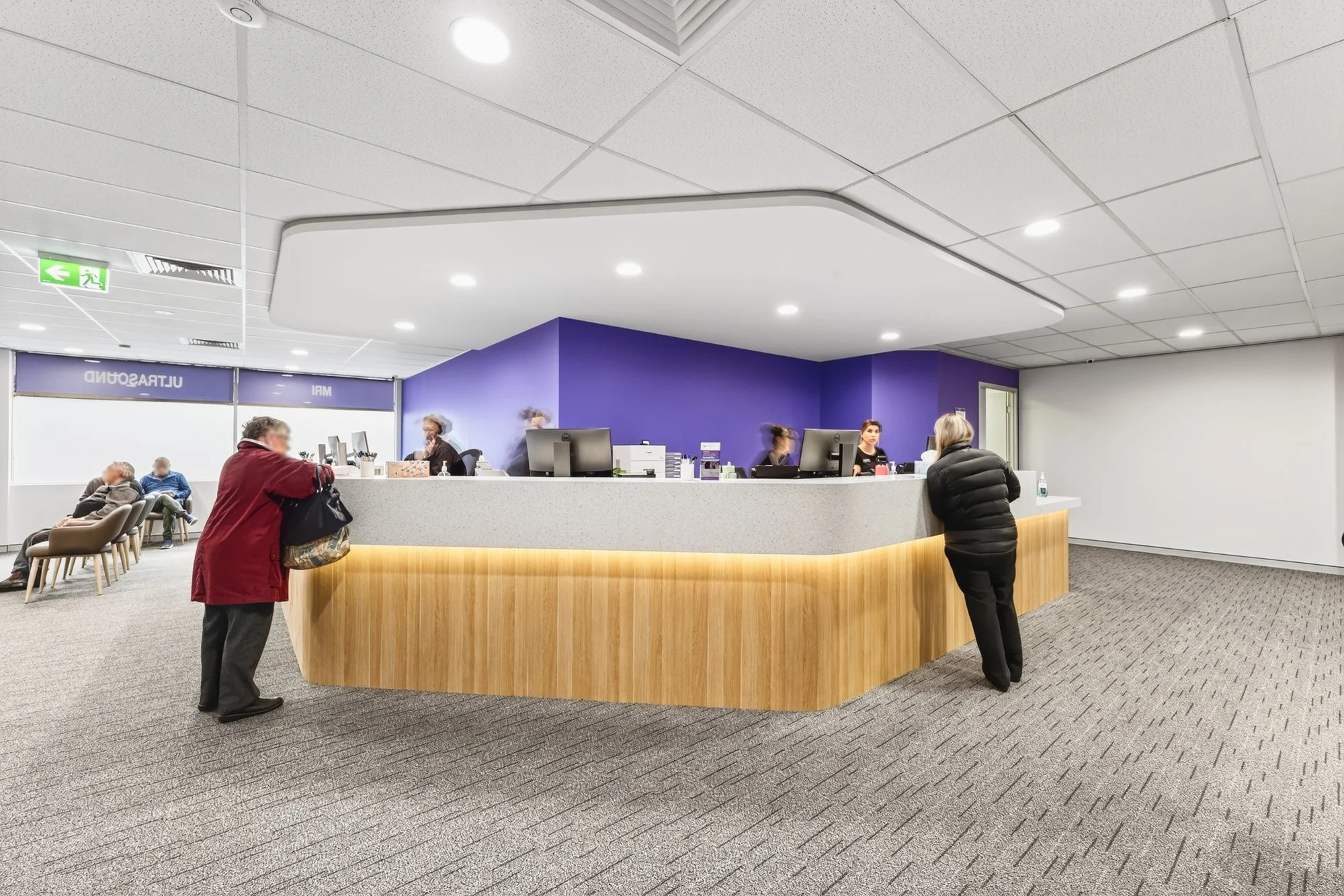 Inside a waiting room with a reception desk. Two women are standing at the desk, and several chairs are occupied by seated individuals in the background. The wall behind the desk is painted purple. The ceiling has recessed lighting.