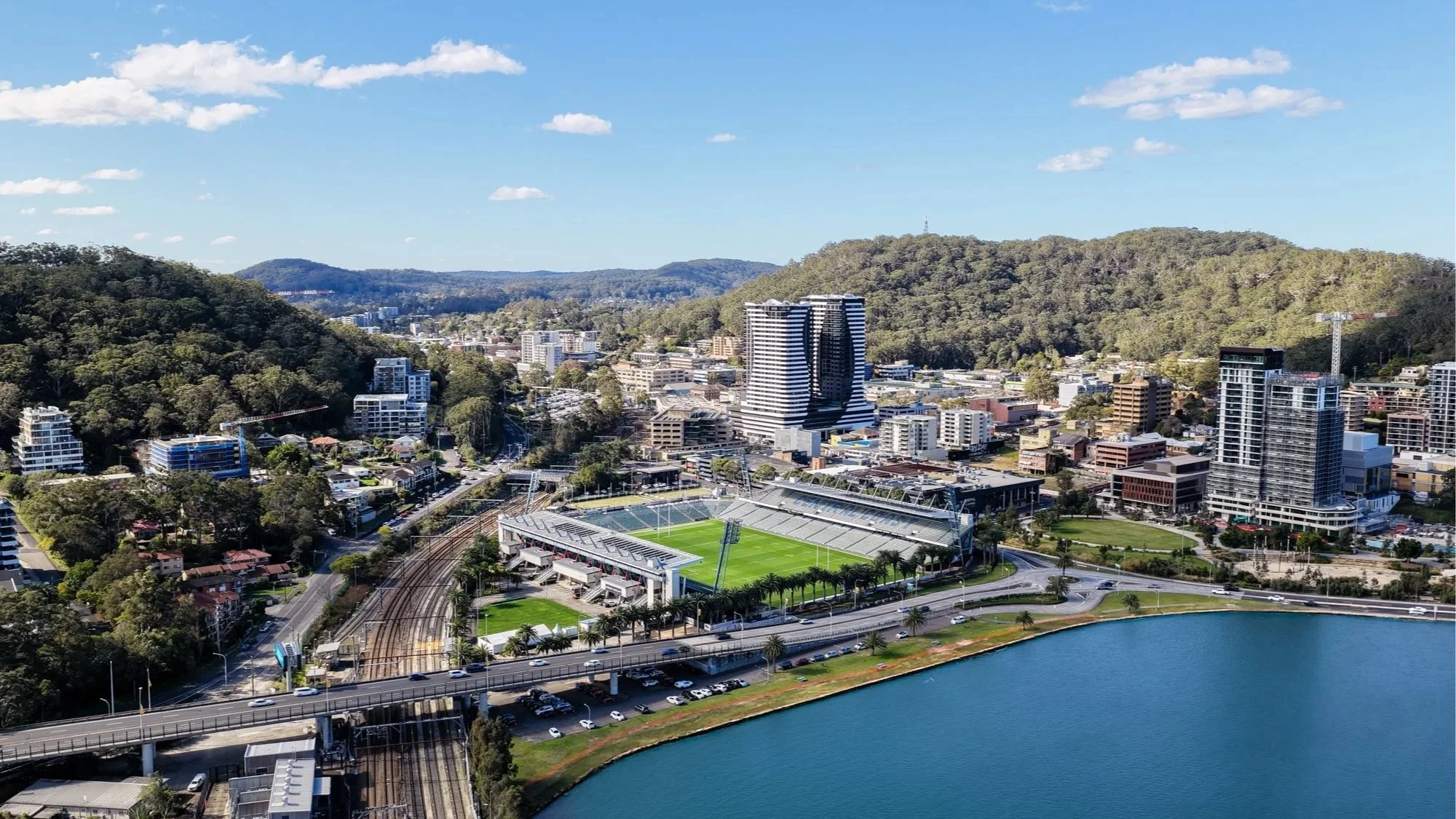 Aerial view of a city with a stadium and a body of water in the foreground, surrounded by trees and hills in the background.