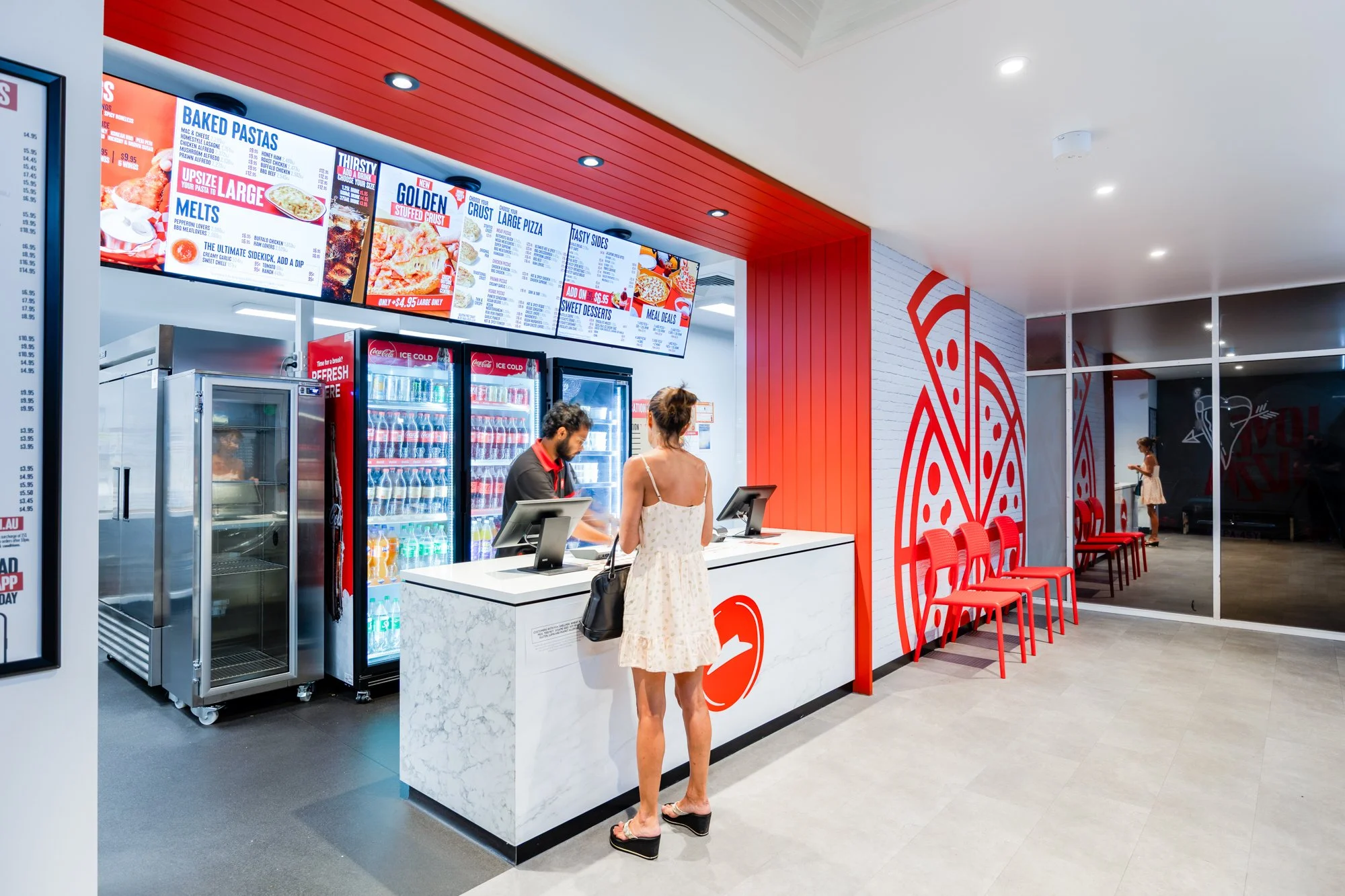 Counter at a fast food restaurant with a woman in a white dress ordering and a staff member taking her order, digital menu screens above displaying food options, red chairs lined against a wall, and a woman in the background looking at her phone.