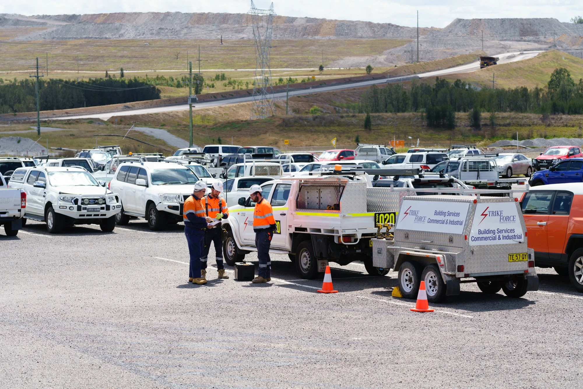 Three workers in orange and blue safety uniforms and white helmets working near a white utility truck and trailer in a large parking lot filled with parked cars, with a hilly landscape and power lines in the background.