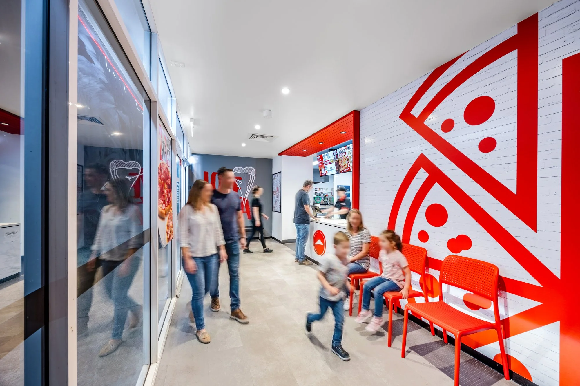 Inside a pizza restaurant, with a white brick wall decorated with a large red pizza slice graphic, red chairs, and a counter where customers order food. Several people are seated and standing, including children and adults, with some digital menu boa