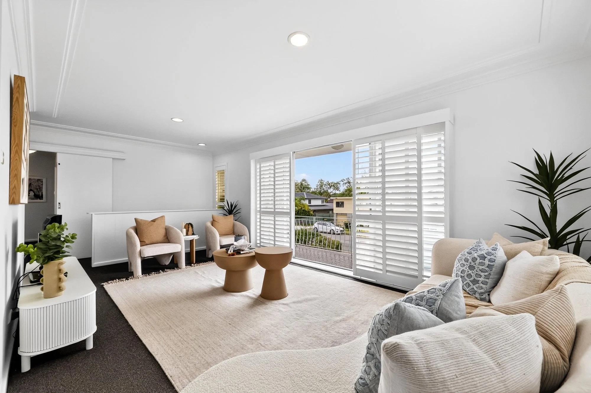 Bright living room with large sliding glass doors, white walls, cream-colored sofa with pillows, two beige armchairs, potted plants, and a beige rug.