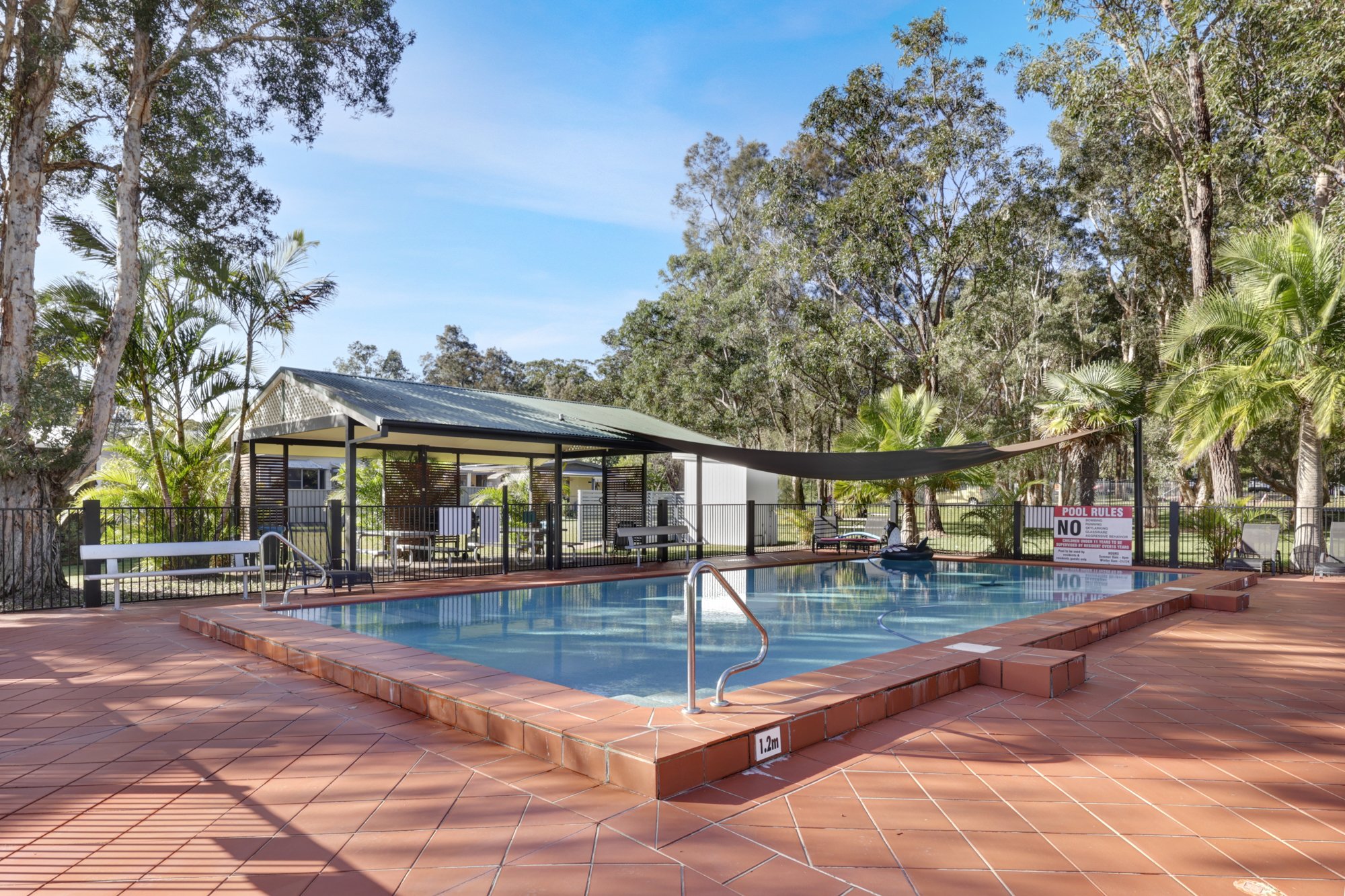 Empty outdoor swimming pool with a terracotta tiled deck, surrounded by trees and a black fence, with pool rules sign and shade structure in the background.