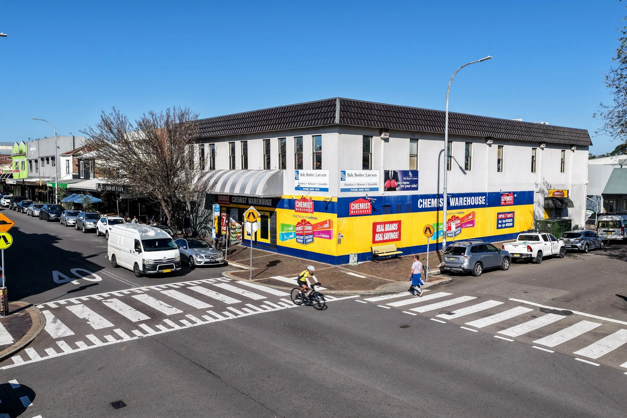 Street corner with parked cars, pedestrians, and a building with commercial signs. Pedestrians cross the crosswalk; cyclists ride along the street. The storefront displays ads for Chemist Warehouse and a law firm, Bale Bosher Lawyers. A blue sky is o