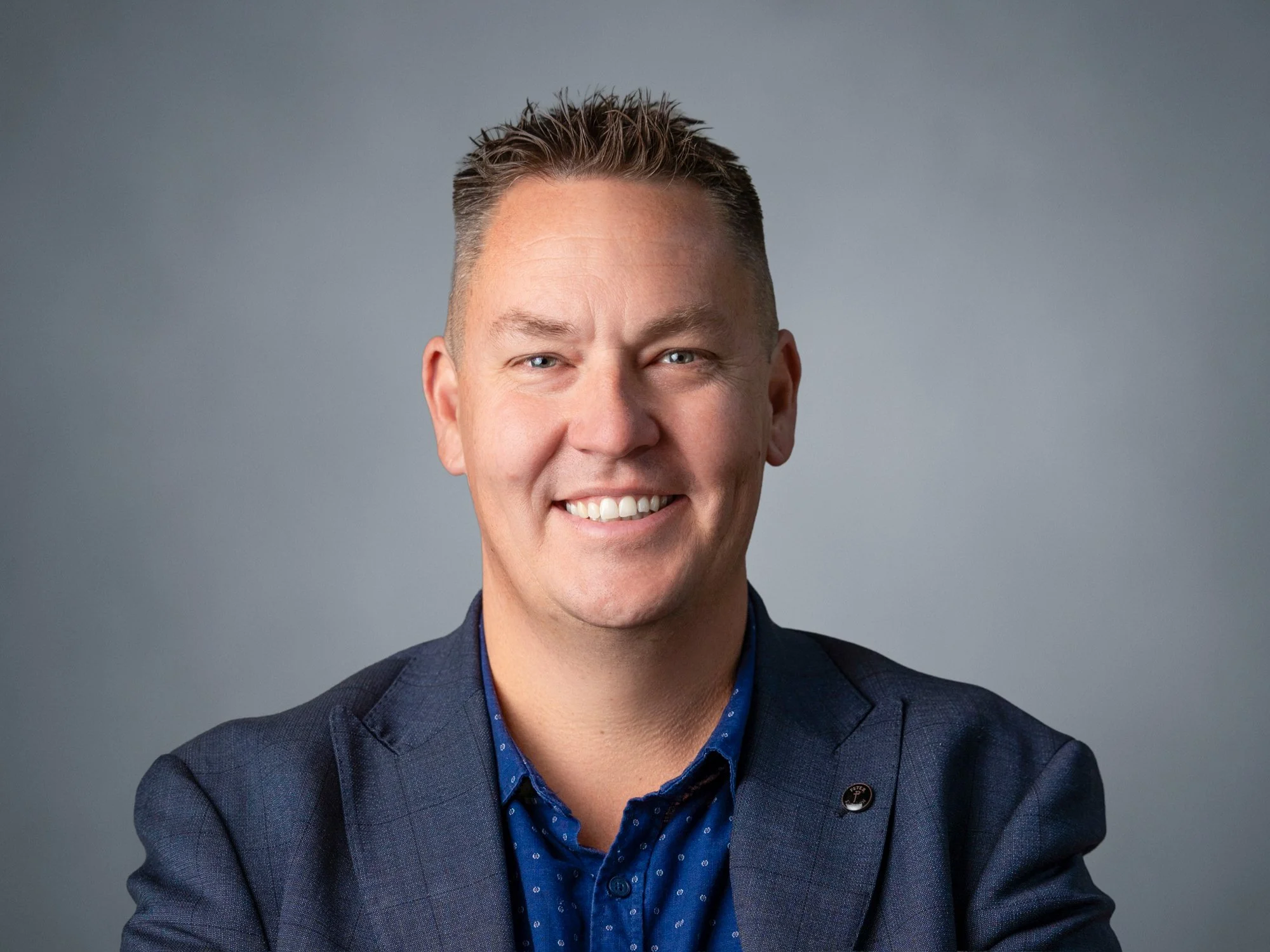 A smiling man wearing a blue blazer and a blue shirt with small white dots, against a plain gray background.