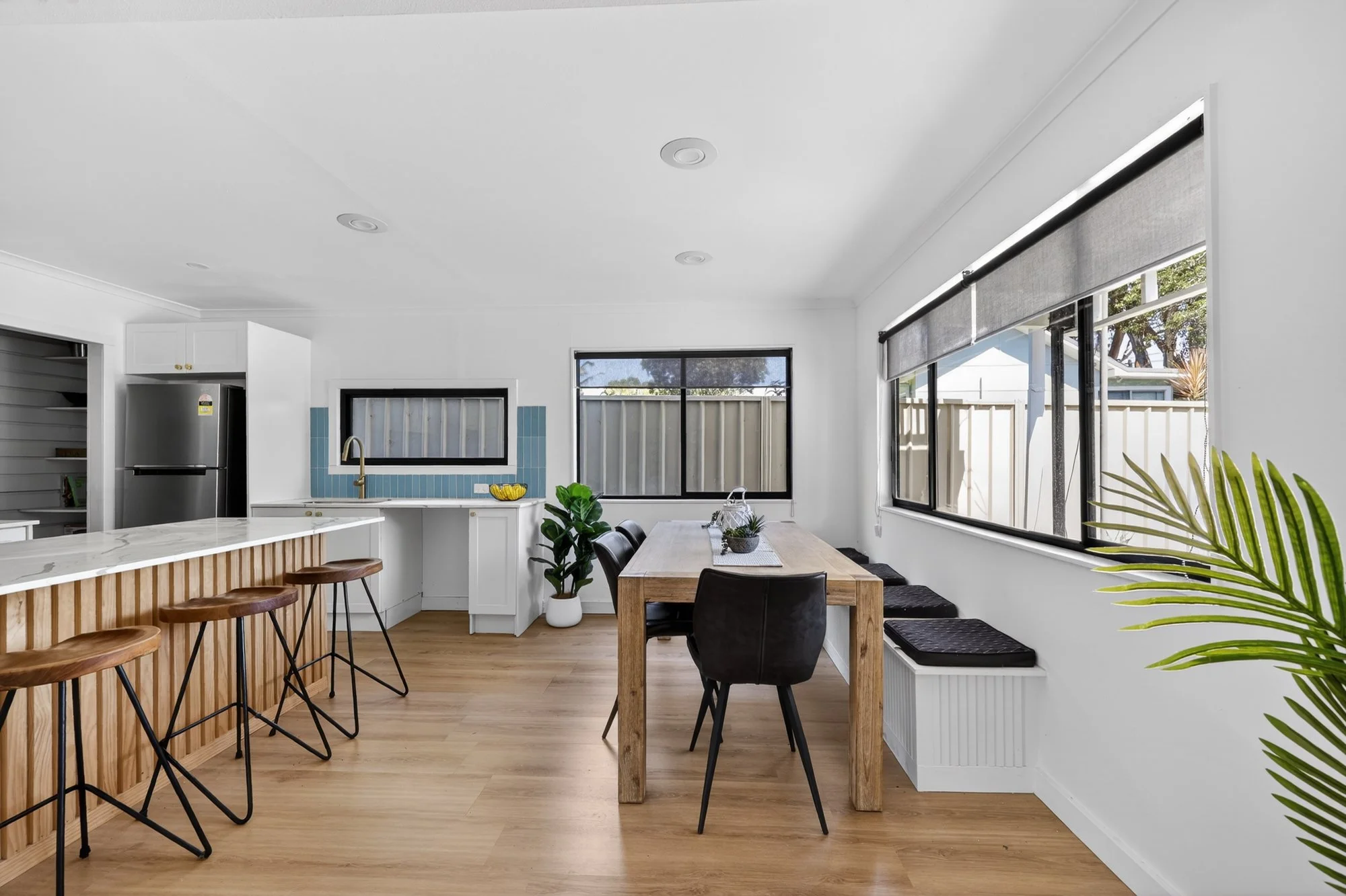 Modern kitchen and dining area with white walls, wooden floors, black framed windows, a wooden dining table with black chairs, a white countertop with bar stools, and some indoor plants.