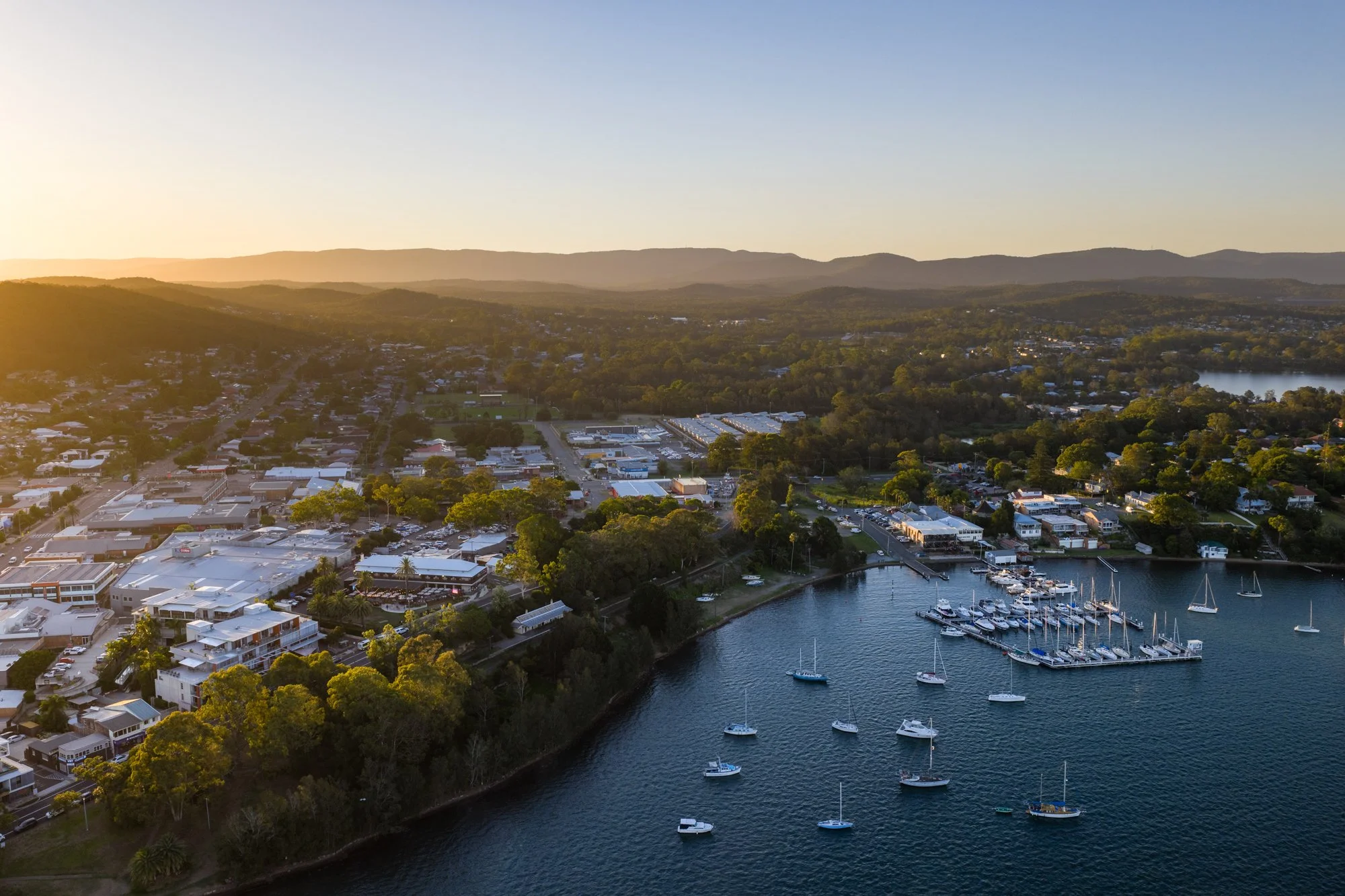 An aerial view of a coastal town at sunset with buildings, trees, and sailboats in the water.