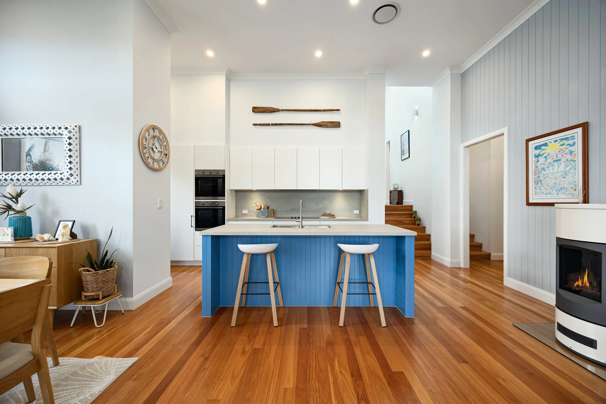 Modern kitchen with blue island, white cabinetry, wooden floor, decorative oars on wall, fireplace, and artwork.