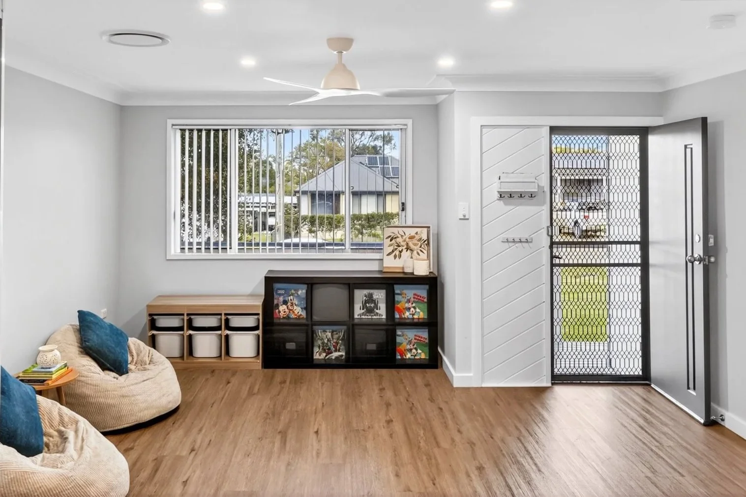 Living room with wooden flooring, white walls, a window with vertical blinds, a door with a security screen, a ceiling fan, built-in storage with baskets, bean bag chairs with blue pillows, and a small bookshelf.