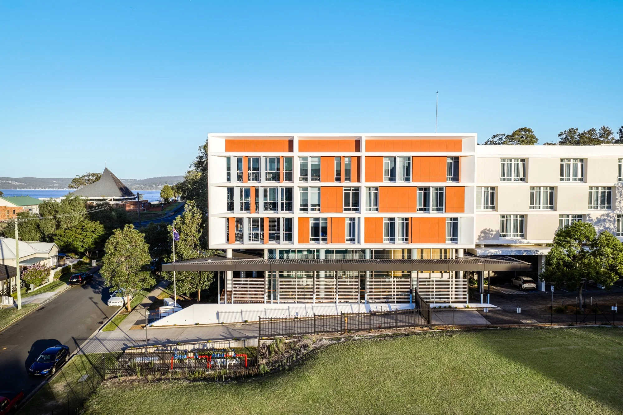 Modern multi-story commercial building with white and orange exterior, large windows, and a covered parking area below. Surrounding greenery and nearby ocean views in the background.