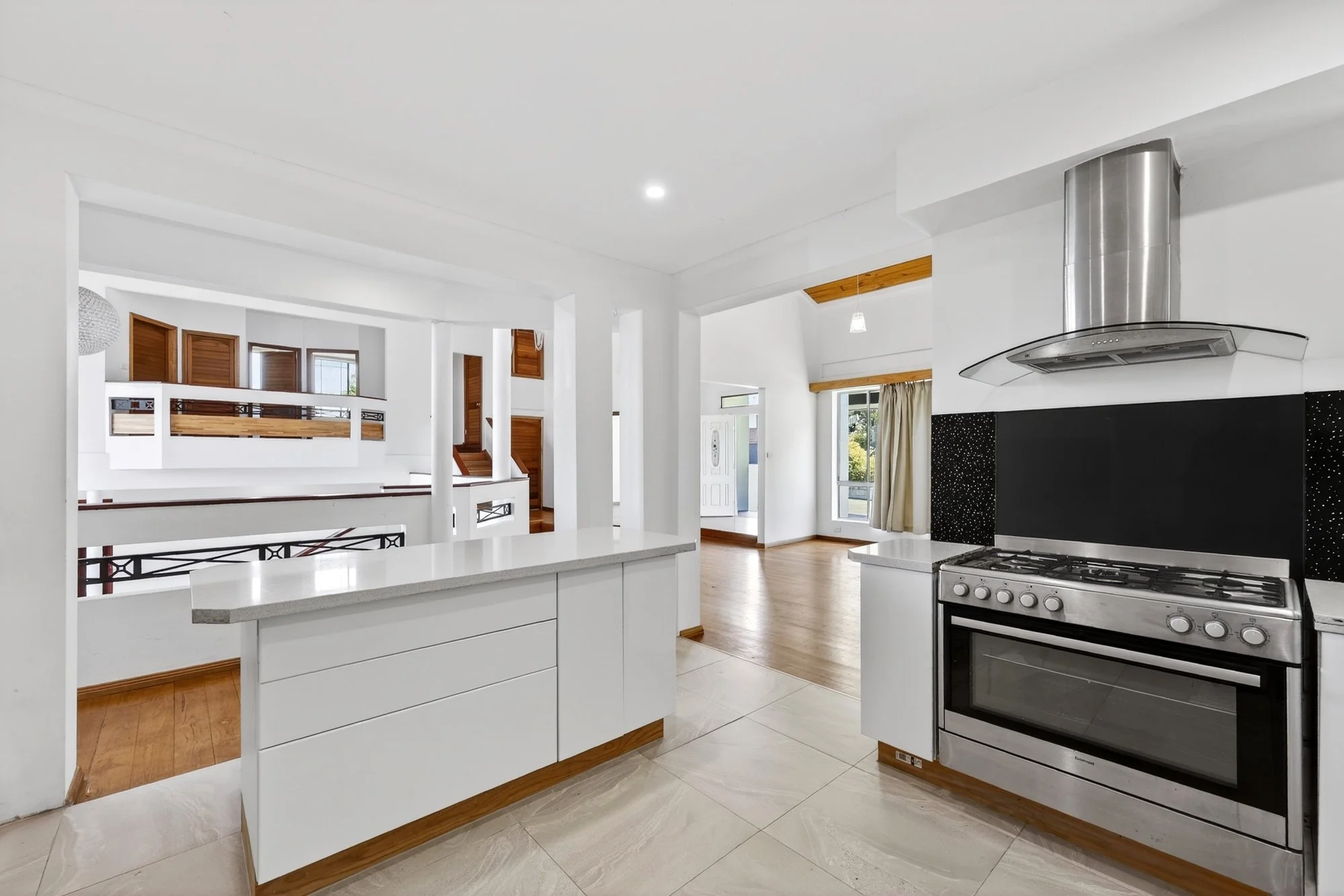 Open-concept kitchen with white cabinetry, a stainless steel stove and oven, black tile backsplash, and a stainless steel range hood. Adjacent living space with wooden flooring and large windows with curtains, leading to an entrance door.