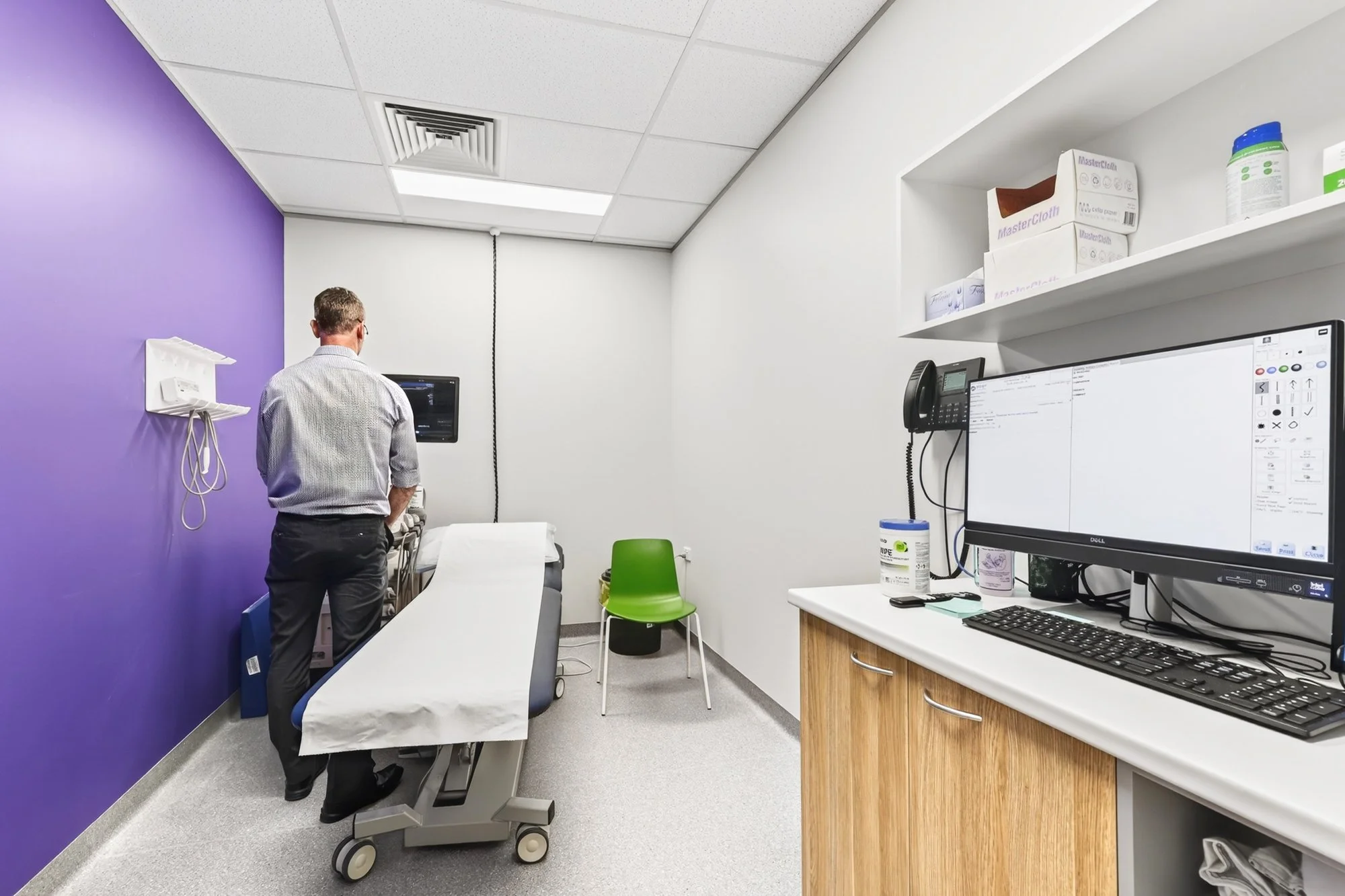 Medical examination room with a doctor standing next to an examination table, a green chair, and medical supplies on a counter, with a purple accent wall and a computer monitor.